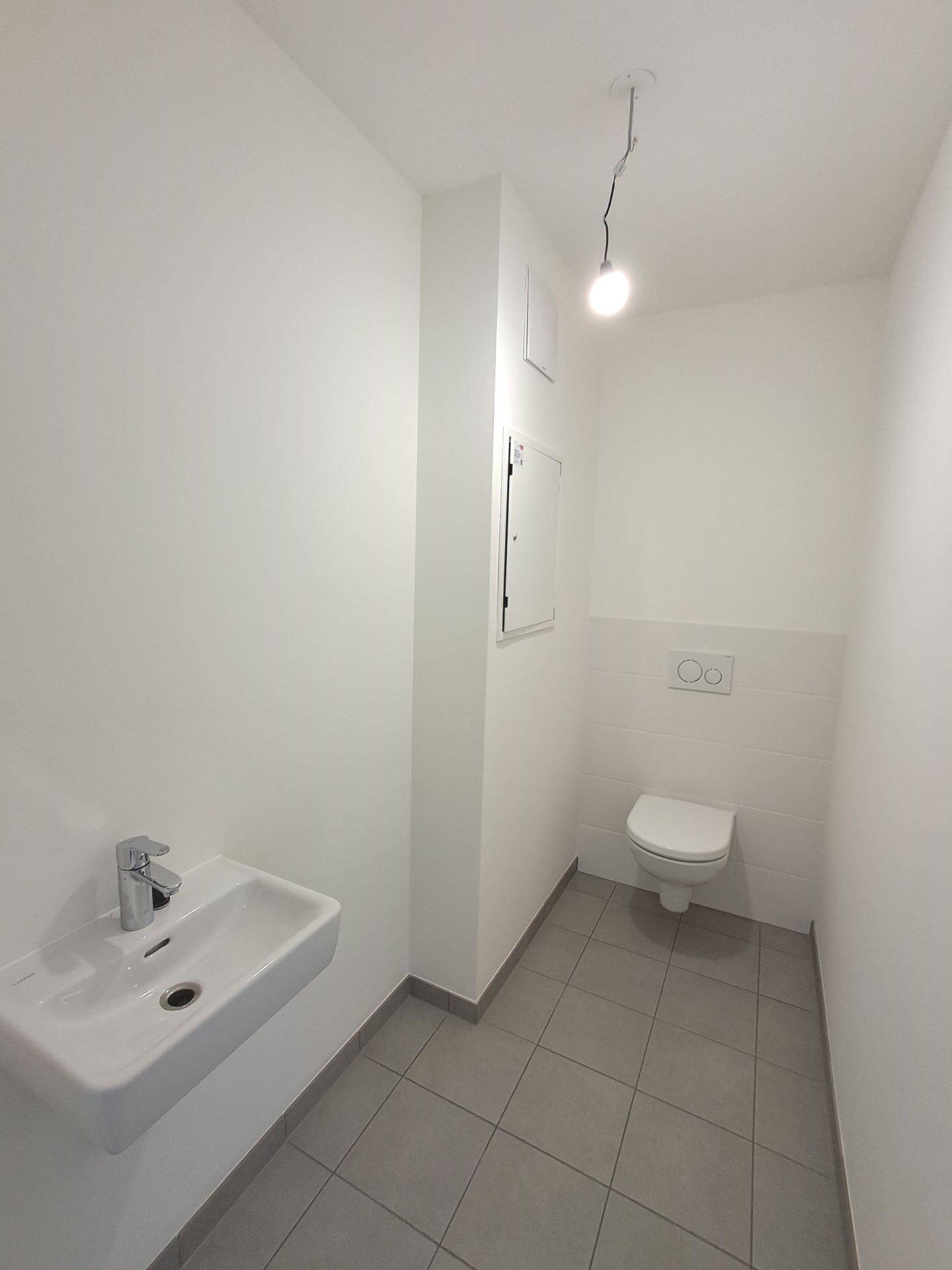 Separates WC mit wandhängender Toilette, kleinem Waschbecken und hellen Fliesen.