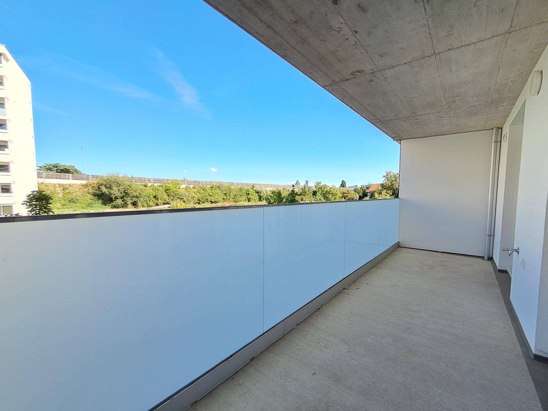 Geräumiger Balkon mit Betonboden und Blick ins Grüne unter blauem Himmel.