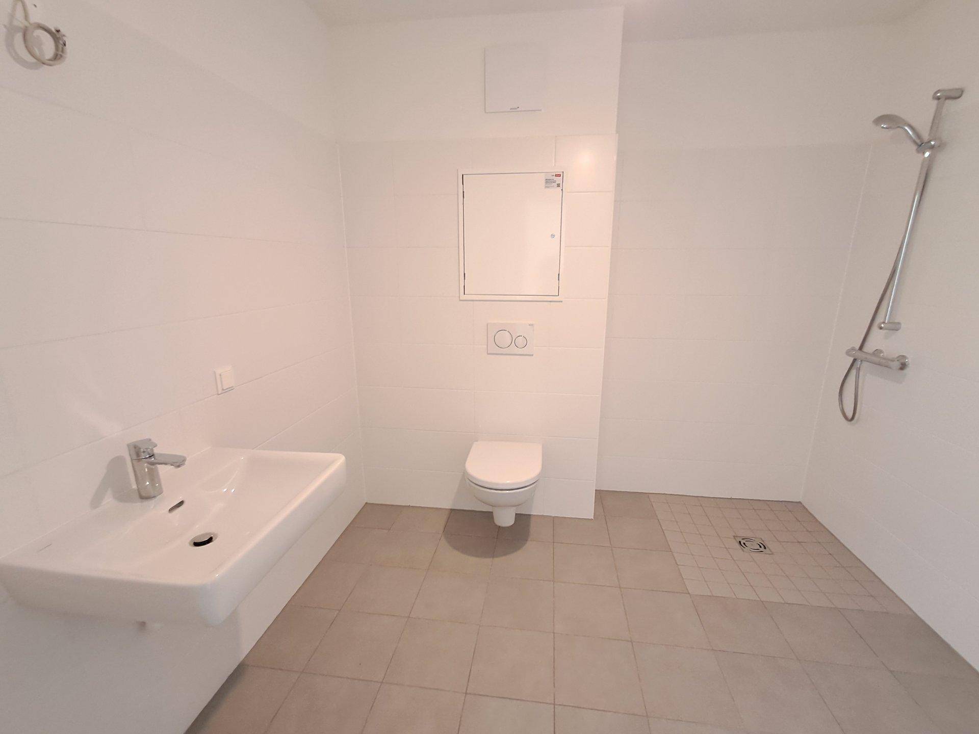Helles Badezimmer mit weißen Wandfliesen, grauen Bodenfliesen, Einzelwaschbecken und wandhängendem WC.