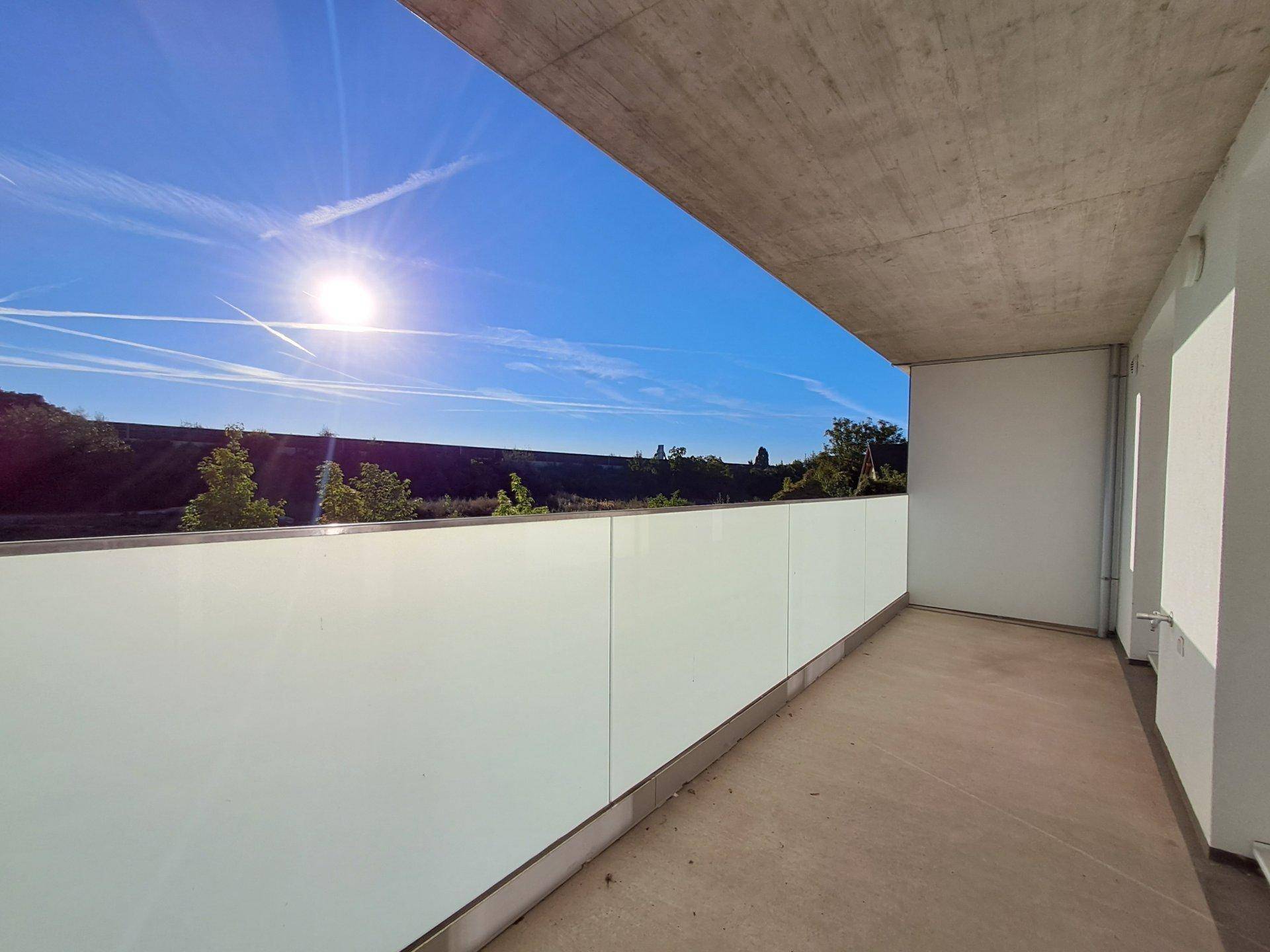 Heller Balkon mit Glasbrüstung und weitem Blick ins Grüne an einem sonnigen Tag.