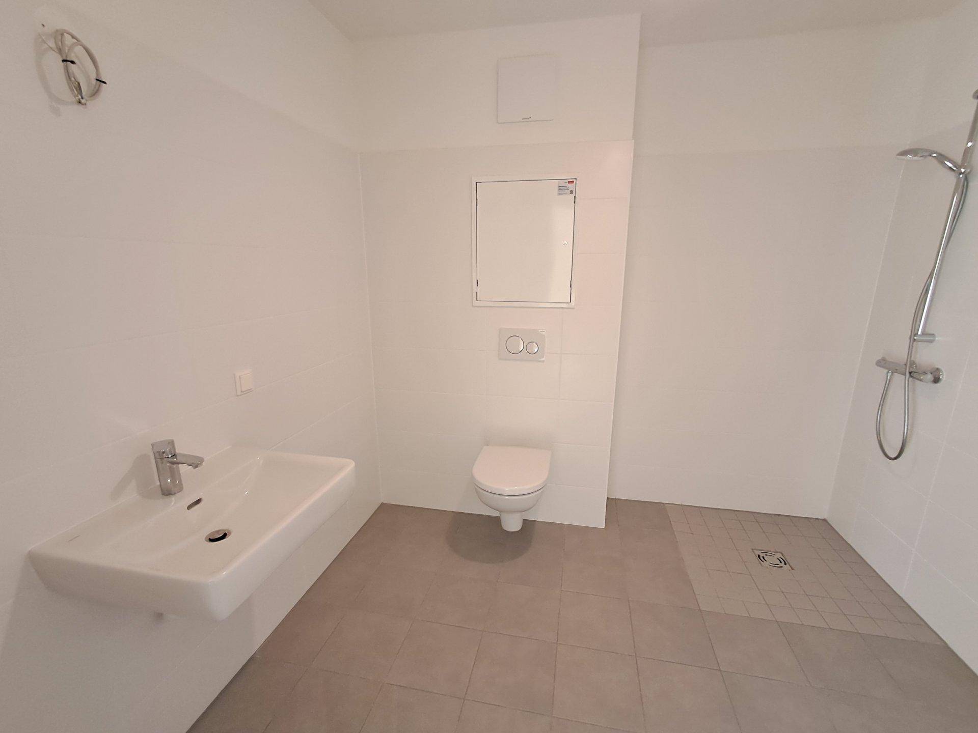 Helles Badezimmer mit Fliesenboden, Wand-WC und Duschbereich, unmöbliert.