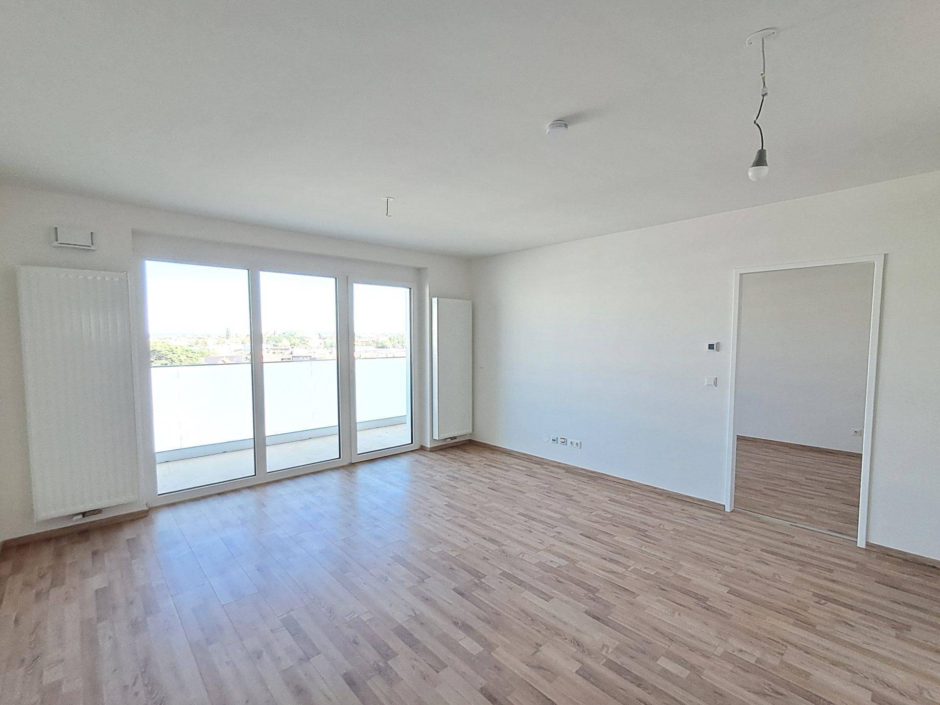 Heller Wohnraum mit großen Balkontüren, Laminatboden und Blick nach draußen.