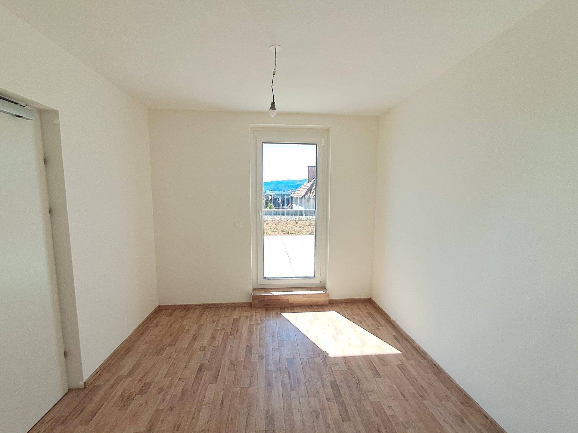 Helles Zimmer mit Laminatboden und einer Tür, die zu einem Außenbereich führt.