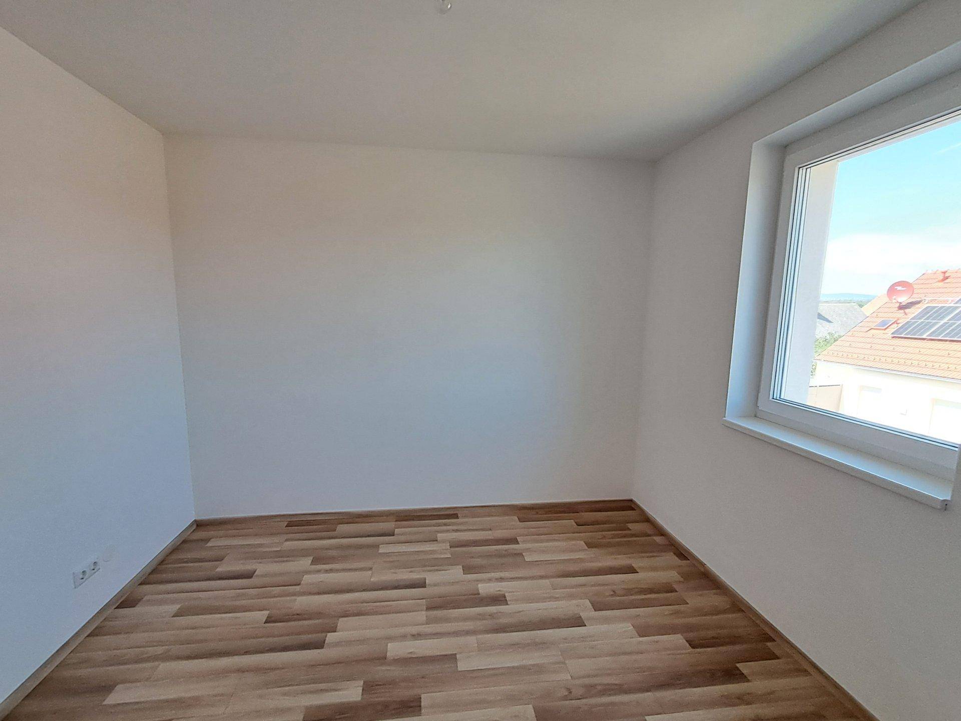 Helles, leeres Schlafzimmer mit Laminatboden und einem Fenster mit Blick auf die Dächer.