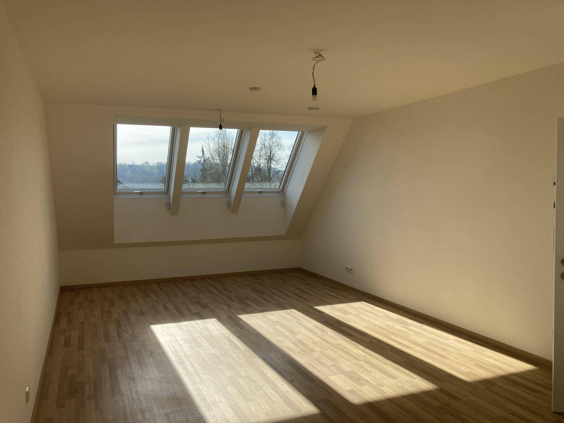 Heller Raum mit Laminatboden, Dachschrägen und großen Dachfenstern, durch die Sonnenlicht fällt.