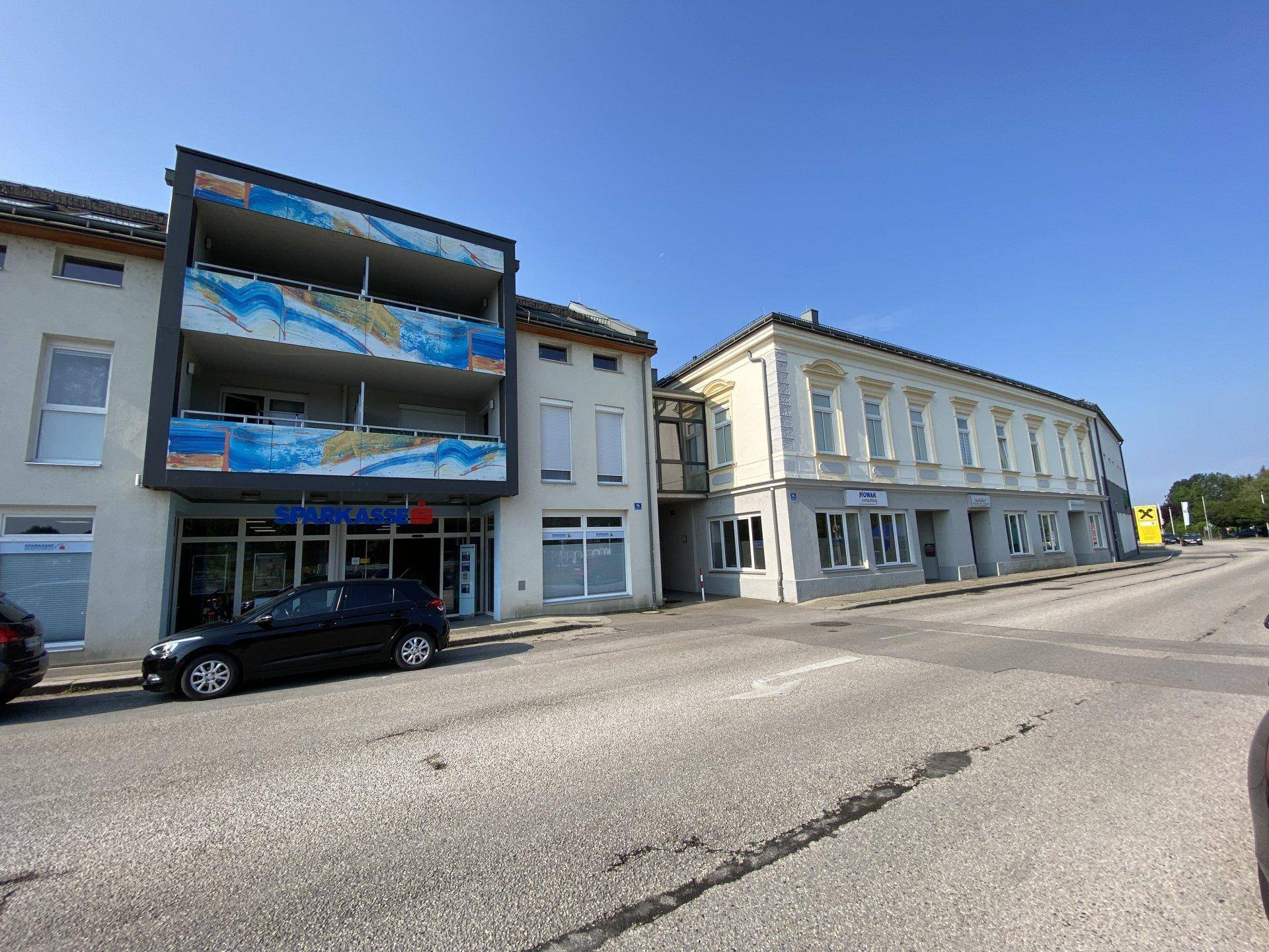 Straßenansicht mit verschiedenen Gebäuden, darunter ein modernes Haus mit farbigen Balkonbrüstungen.