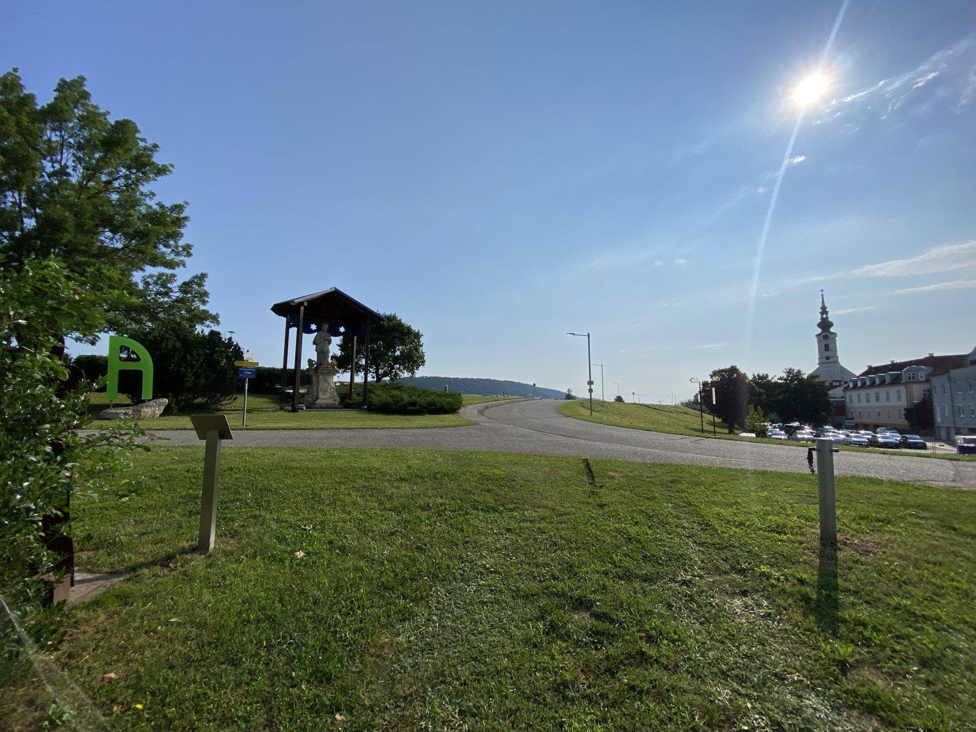 Weitläufige Landschaft mit einer Straße, einem kleinen Schrein und einem Kirchturm in der Ferne.
