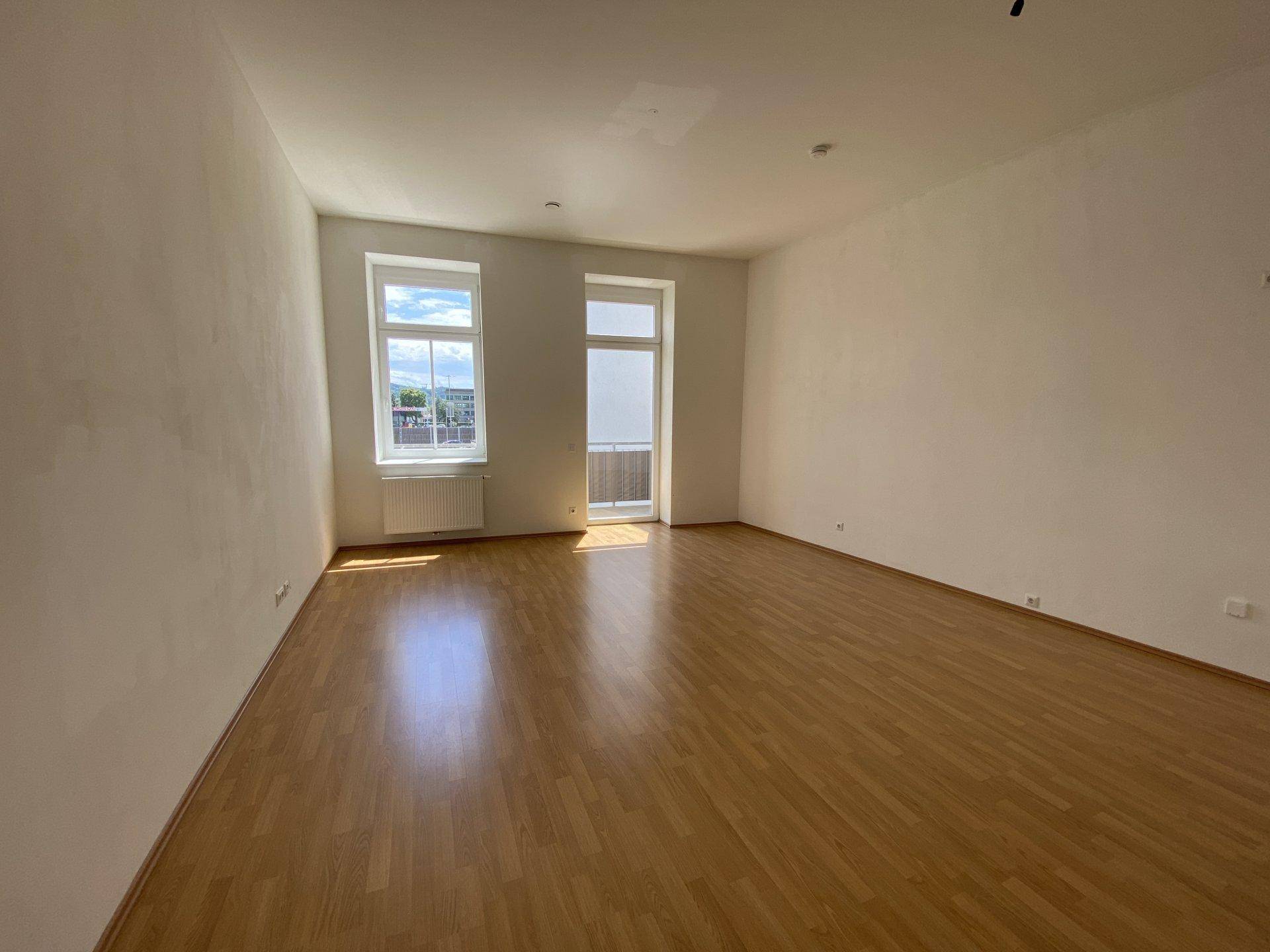 Helles, leeres Zimmer mit Holzboden, großen Fenstern und Zugang zu einem Balkon.