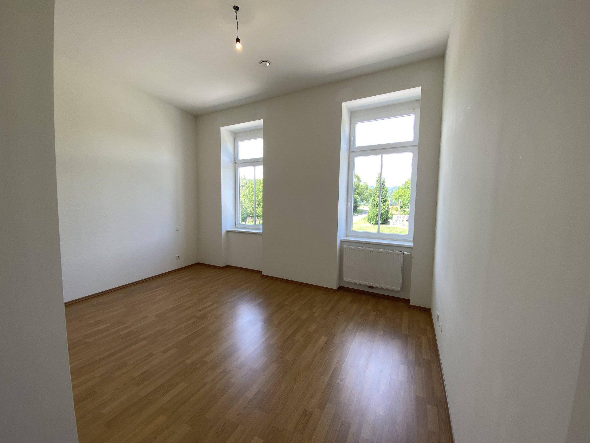 Helles, leeres Zimmer mit Holzboden und zwei Fenstern, die einen grünen Ausblick bieten.
