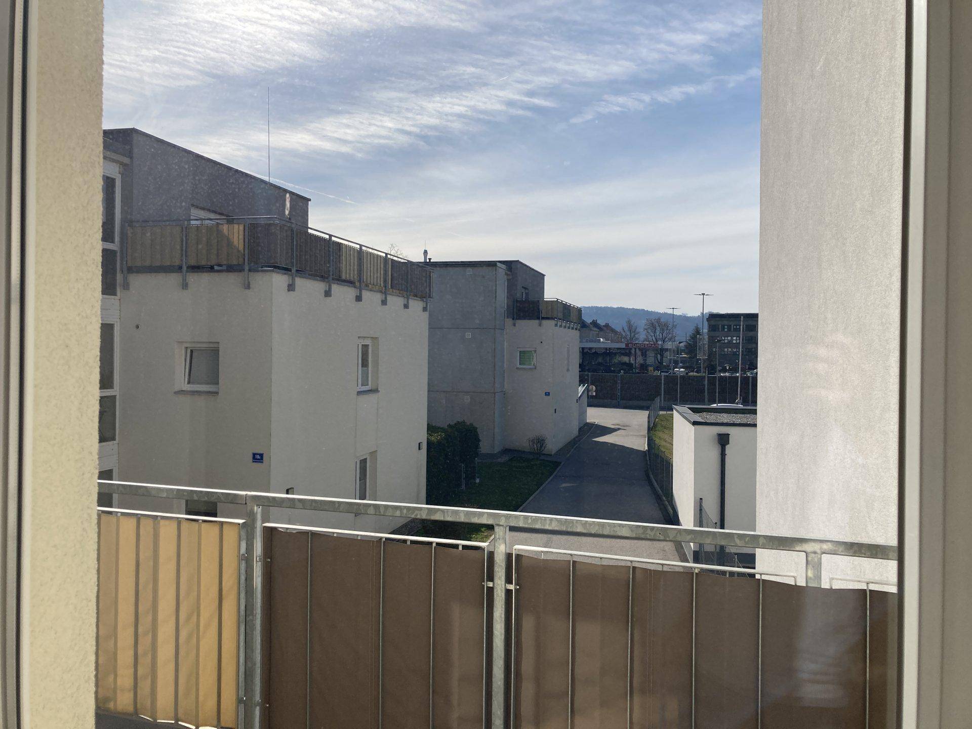 Blick aus einem Fenster oder von einem Balkon auf umliegende Gebäude und eine hügelige Landschaft.