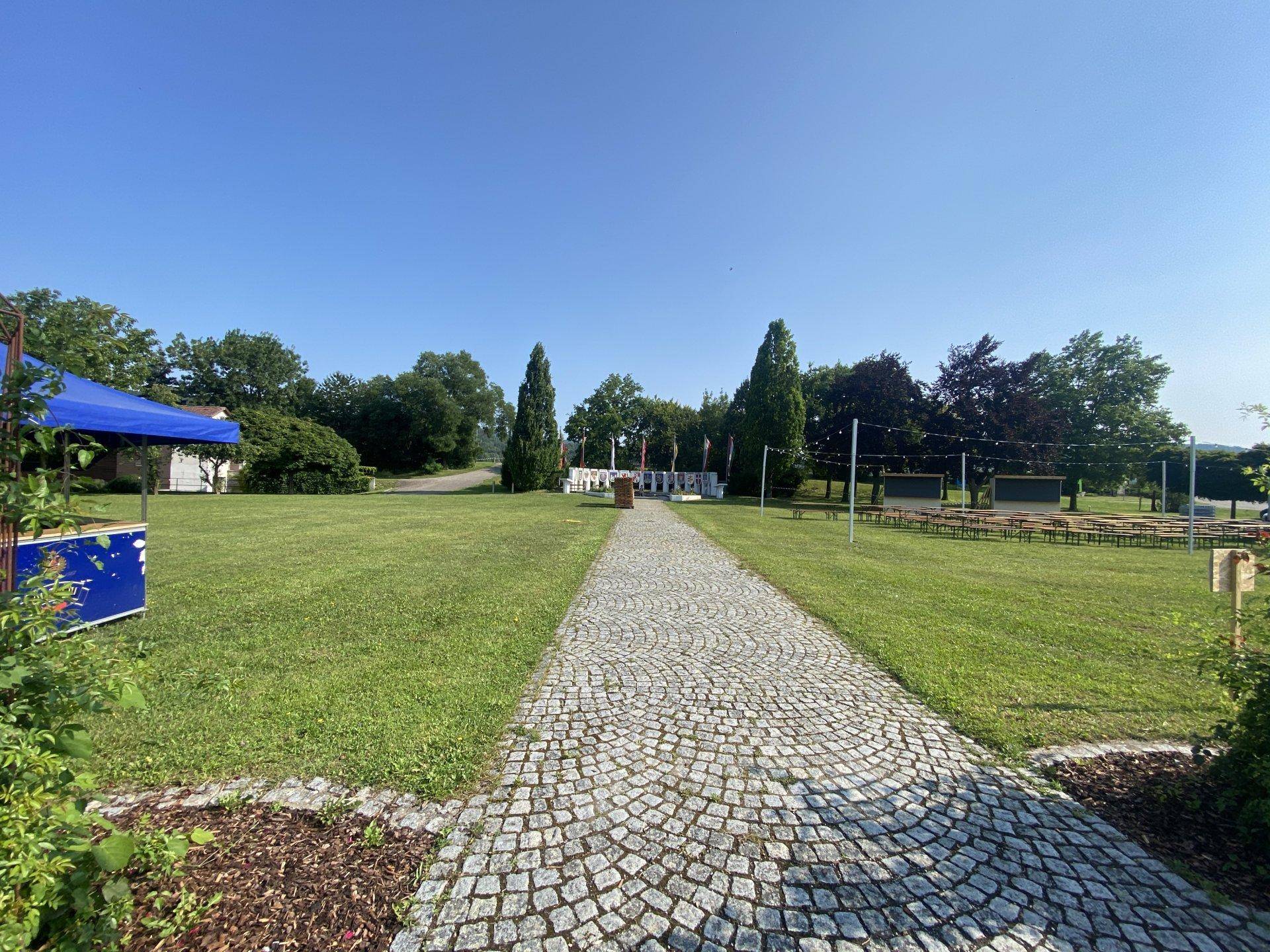 Große Grünfläche mit Kopfsteinpflasterweg, Denkmal und vorbereiteten Sitzgelegenheiten für eine Veranstaltung.