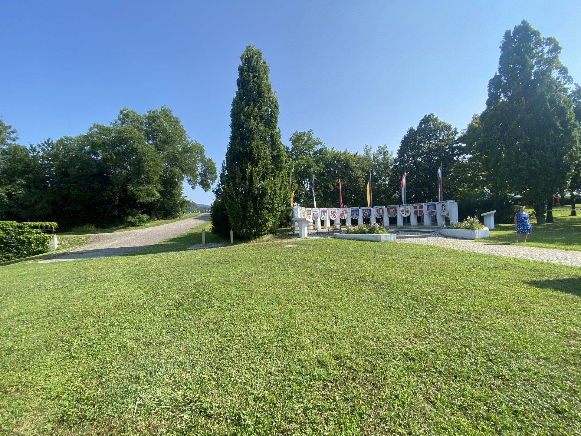 Weitläufige Grünfläche mit einem Denkmal und hohen Bäumen unter strahlend blauem Himmel.