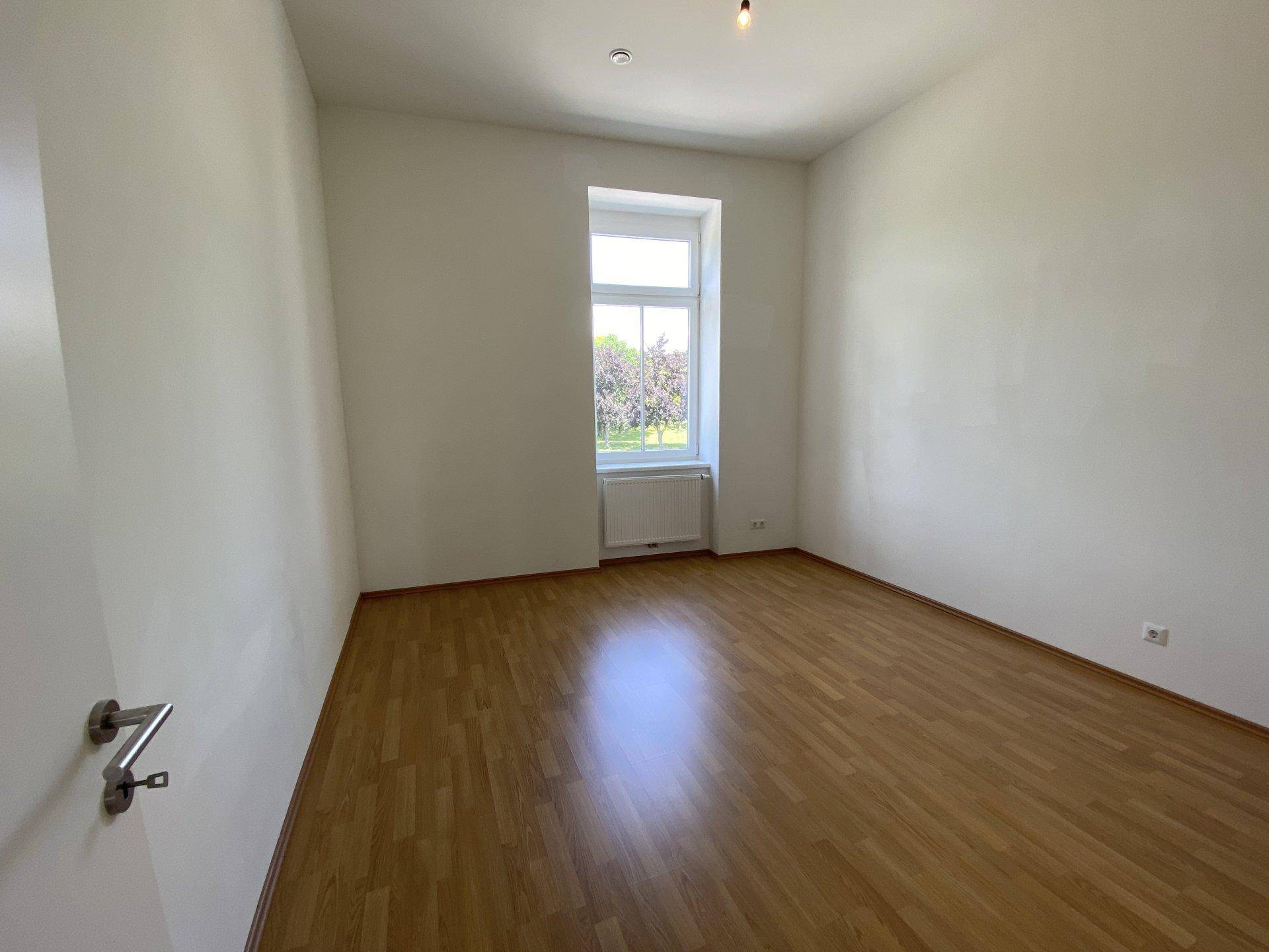 Leeres Zimmer mit Holzboden und einem einzelnen Fenster, das viel Tageslicht hereinlässt.