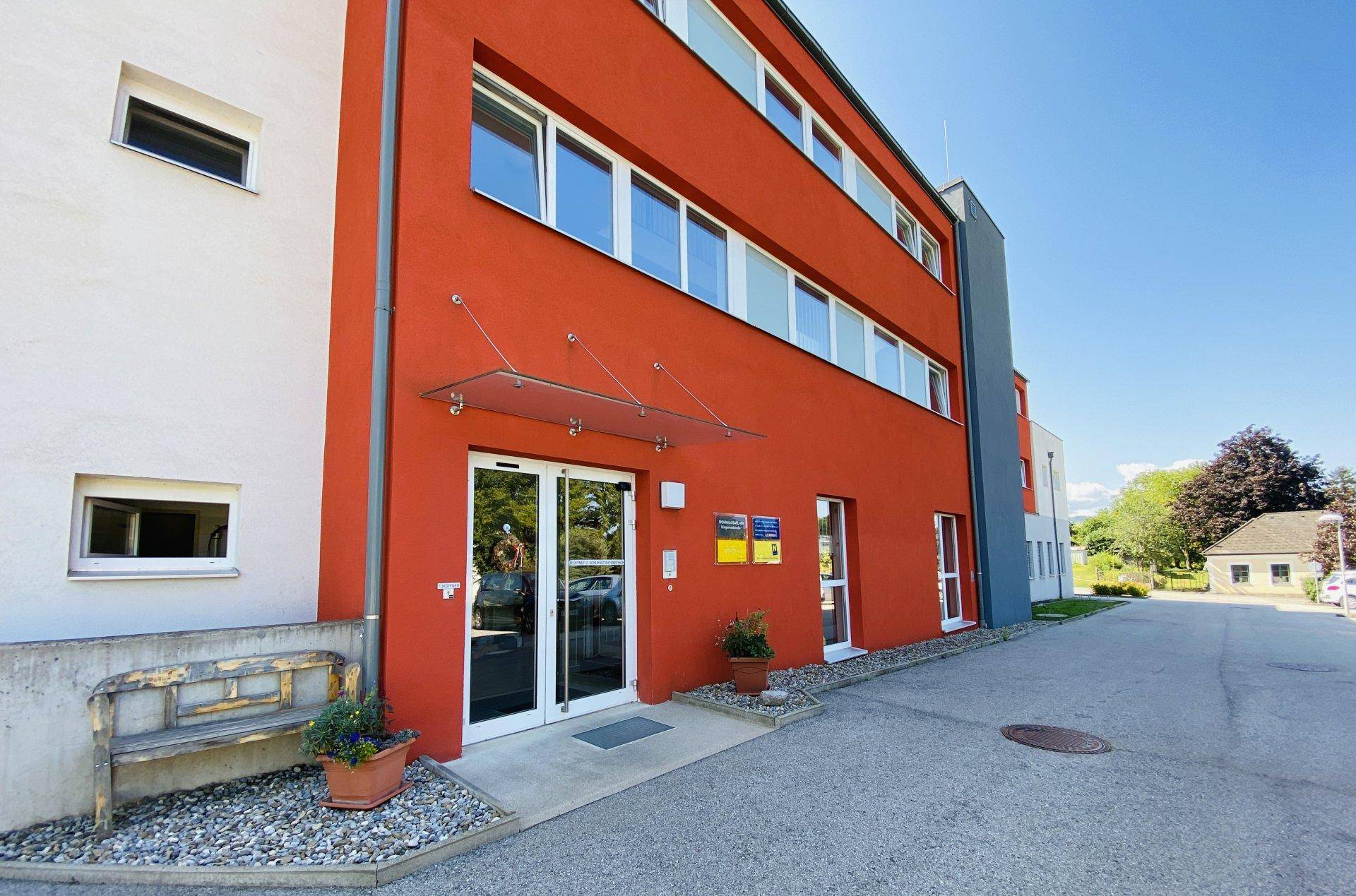 Eingangsbereich eines modernen Gebäudes mit roter Fassade und Glasvordach an einem sonnigen Tag.