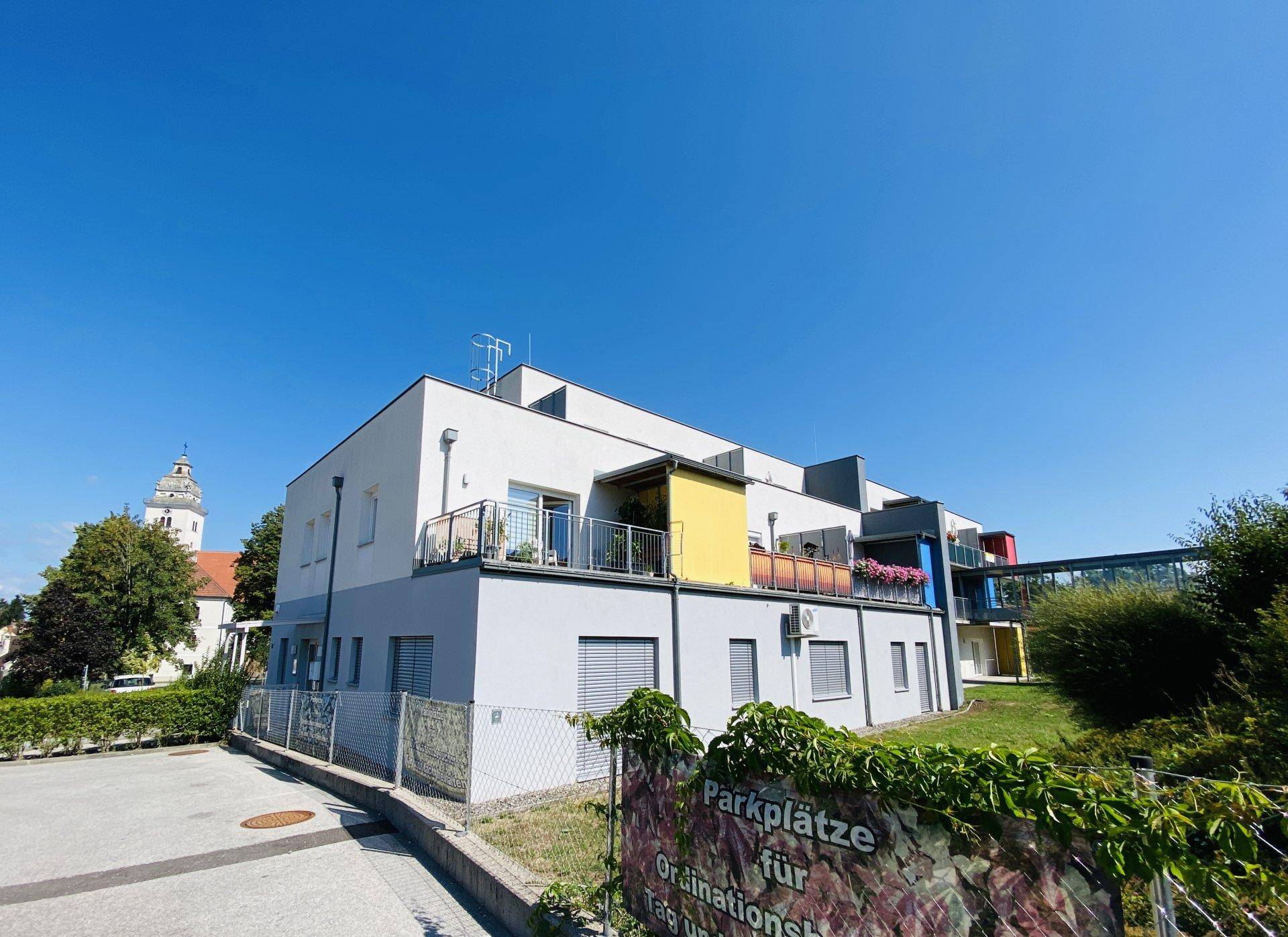 Modernes Gebäude mit Balkonen und Parkplätzen, im Hintergrund ist ein Kirchturm sichtbar.