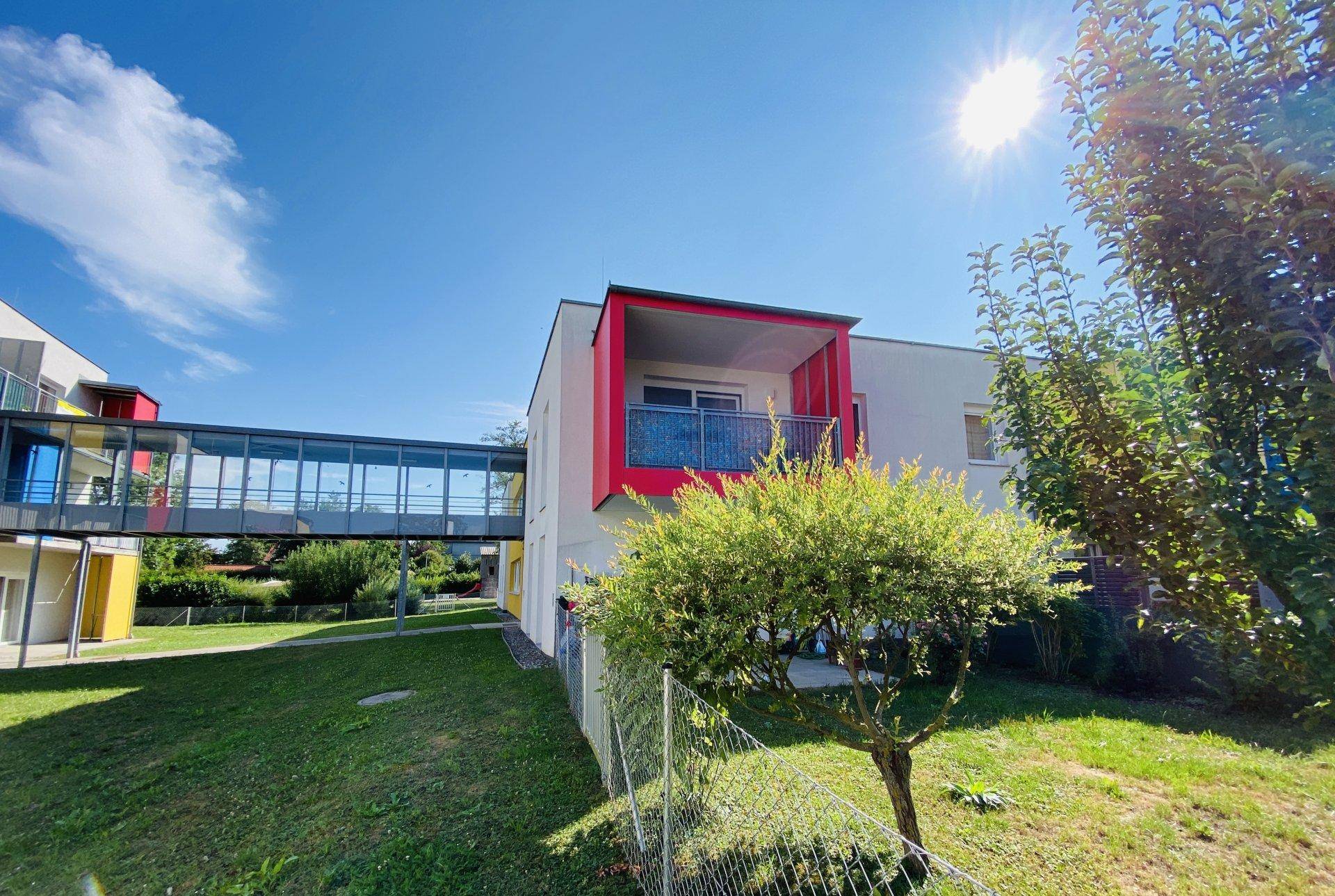 Modernes Gebäude mit rotem Balkon und einer gläsernen Verbindungsbrücke unter strahlendem Sonnenschein.