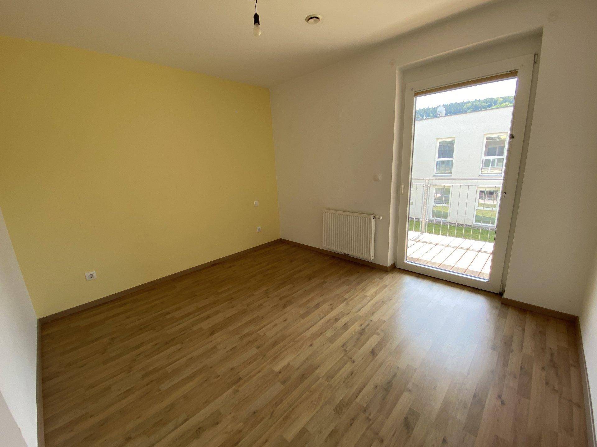 Schlafzimmer mit gelber Akzentwand, Laminatboden und Zugang zu einem Balkon.