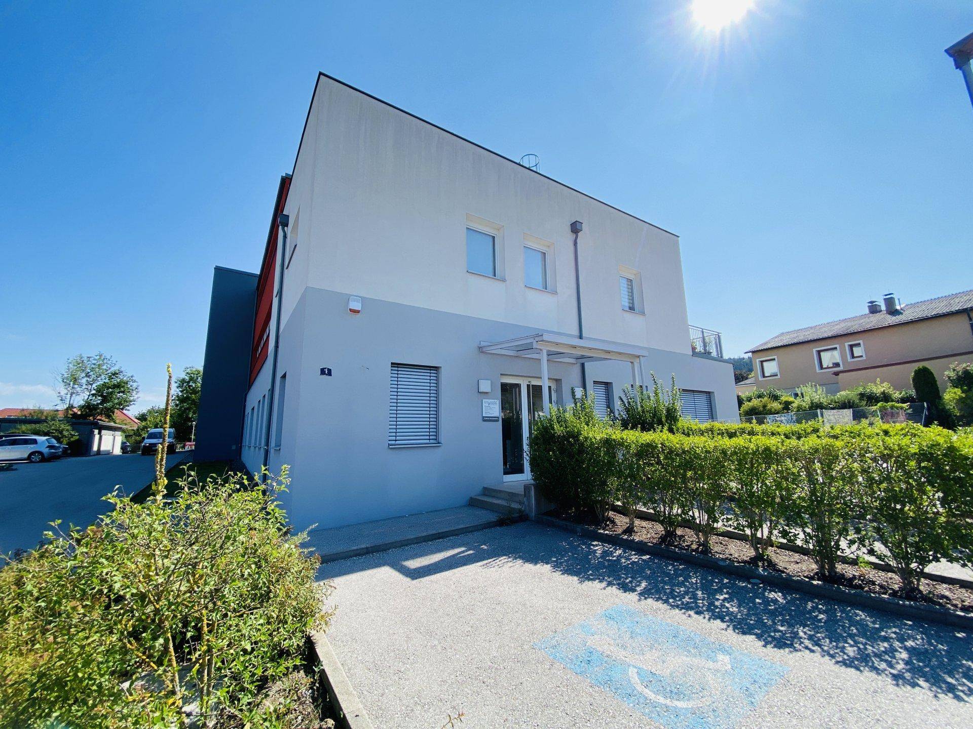 Eingangsbereich eines modernen Gebäudes mit Vordach und Hecke an einem sonnigen Tag.