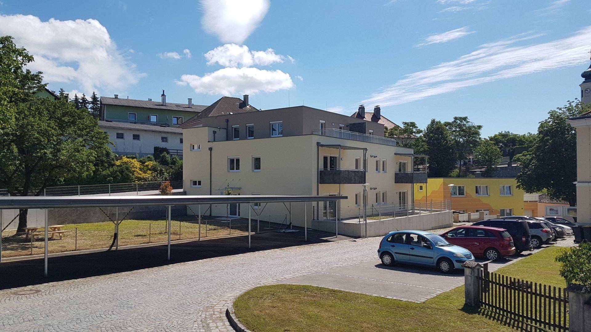 Gesamtansicht des Wohngebäudes mit Parkplätzen, Carport und umliegender Bebauung unter blauem Himmel.