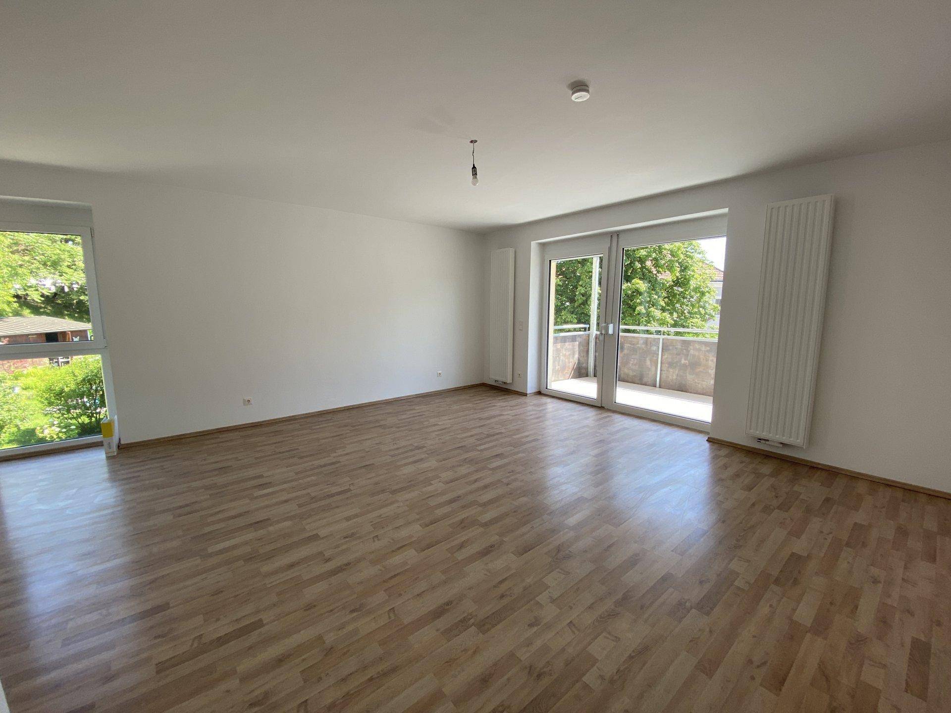 Helles, leeres Wohnzimmer mit Laminatboden, großen Fenstern und Zugang zum Balkon.