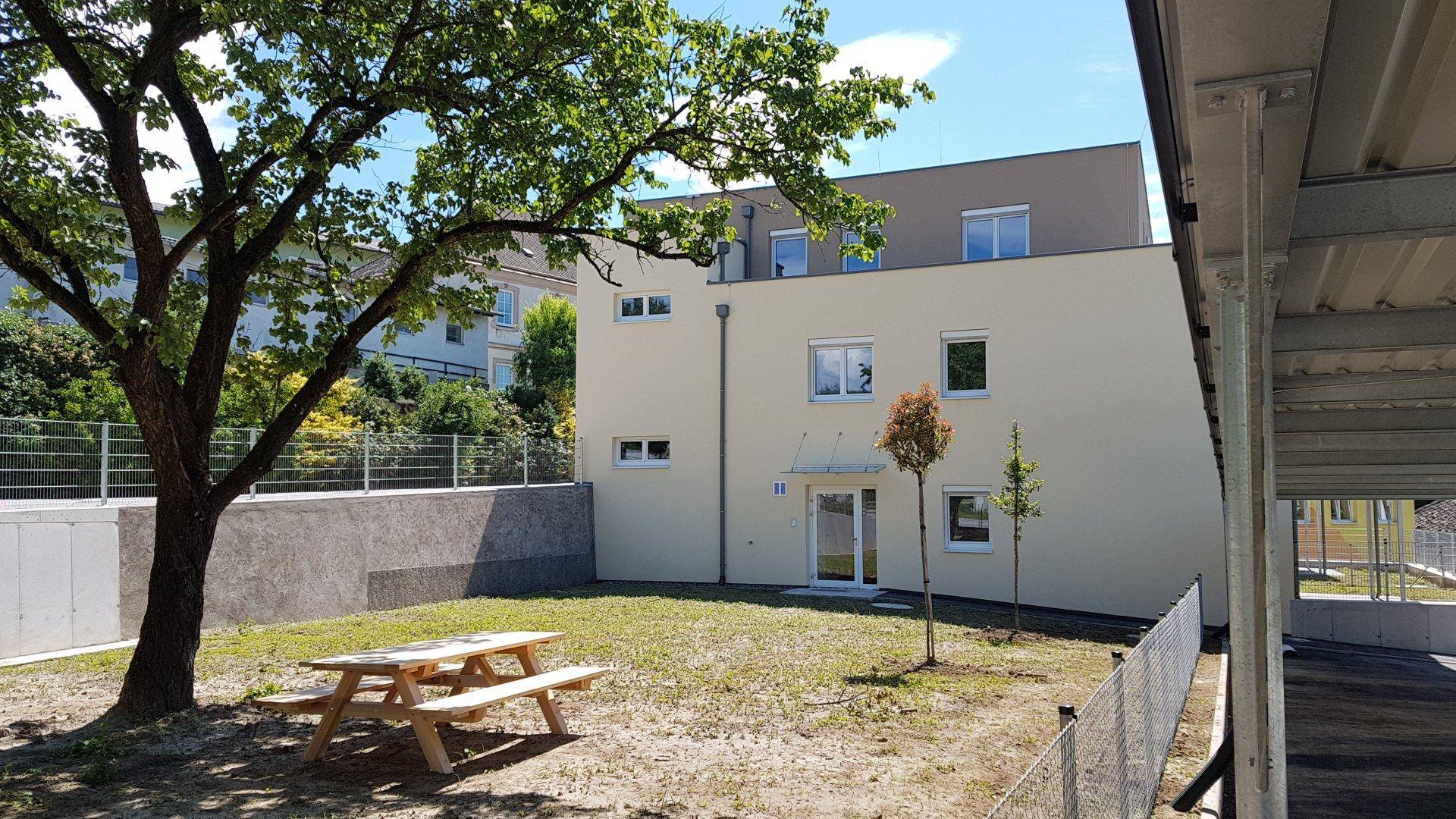 Gemeinschaftsgarten mit Rasenfläche und Picknicktisch, umgeben von Bäumen und dem Gebäude.
