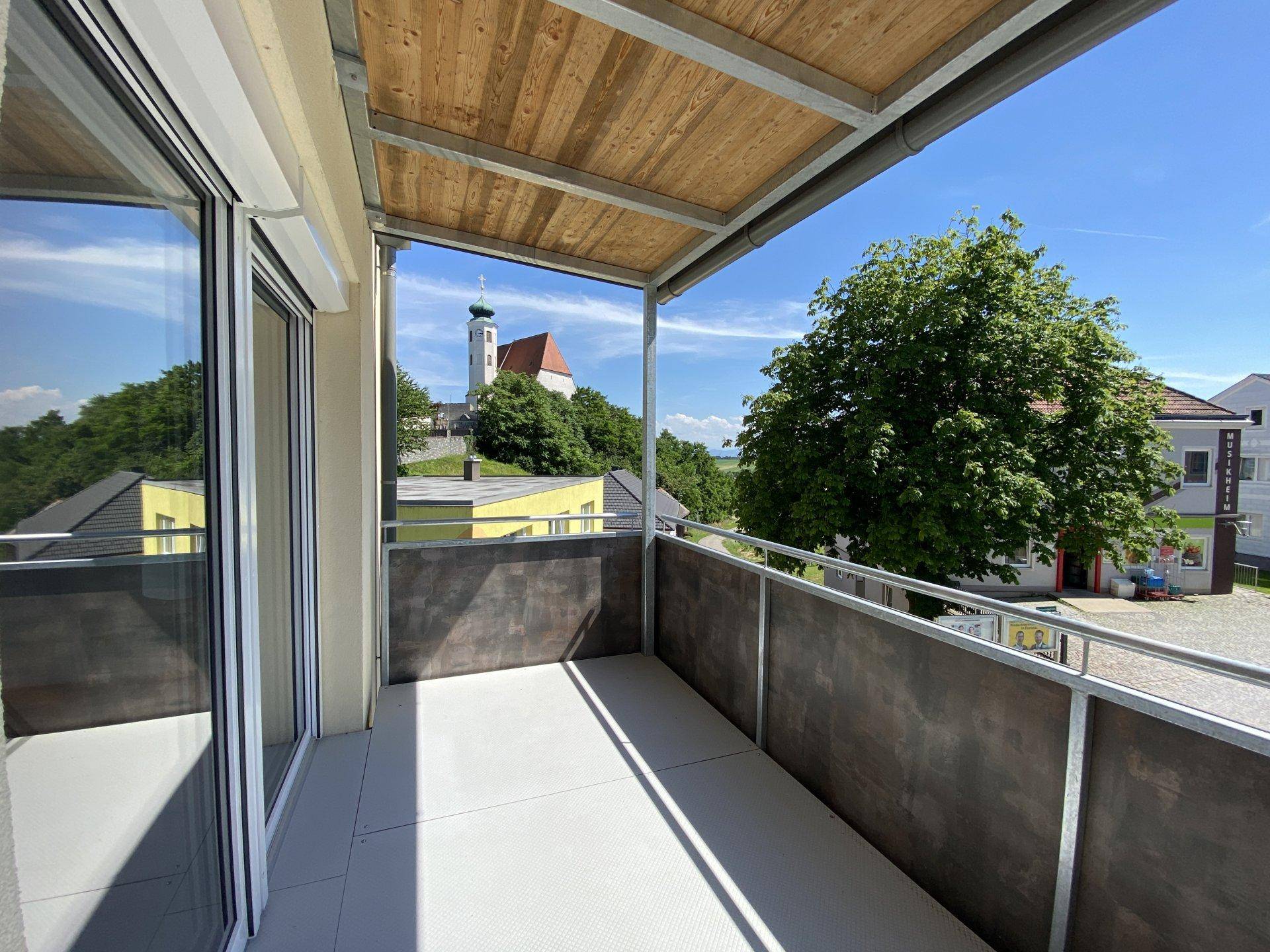 Überdachter Balkon mit Holzdach und Blick auf eine Kirche sowie grüne Umgebung.