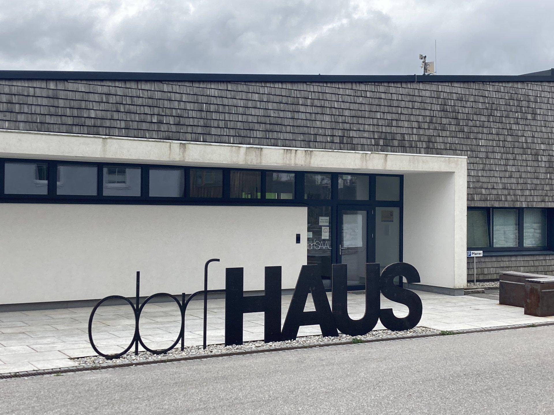 Modernes Dorfhaus mit Schindeldach und großer Beschriftung im Eingangsbereich.