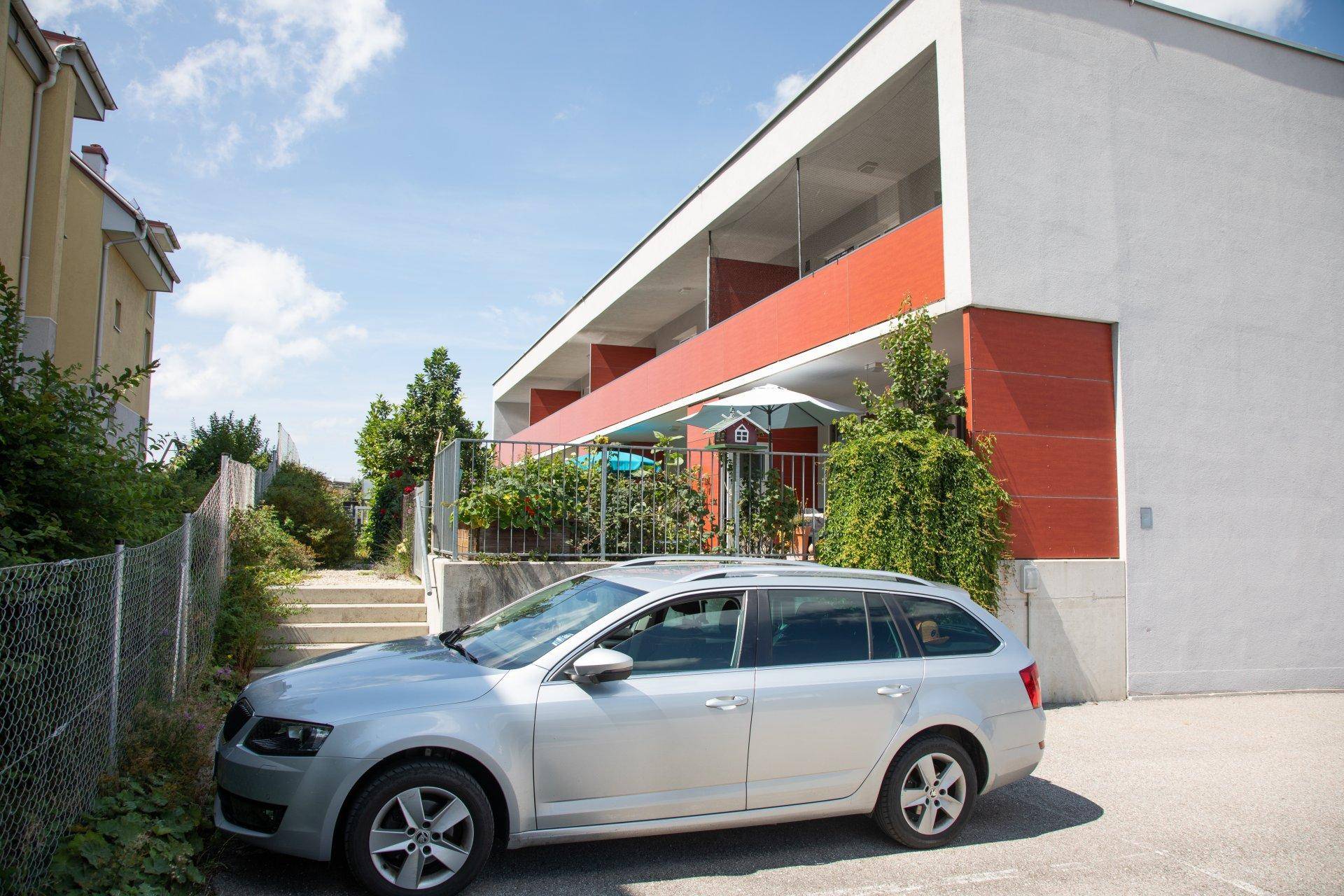 Modernes Wohngebäude mit roten Akzenten und Balkonen, daneben ein geparktes Auto.