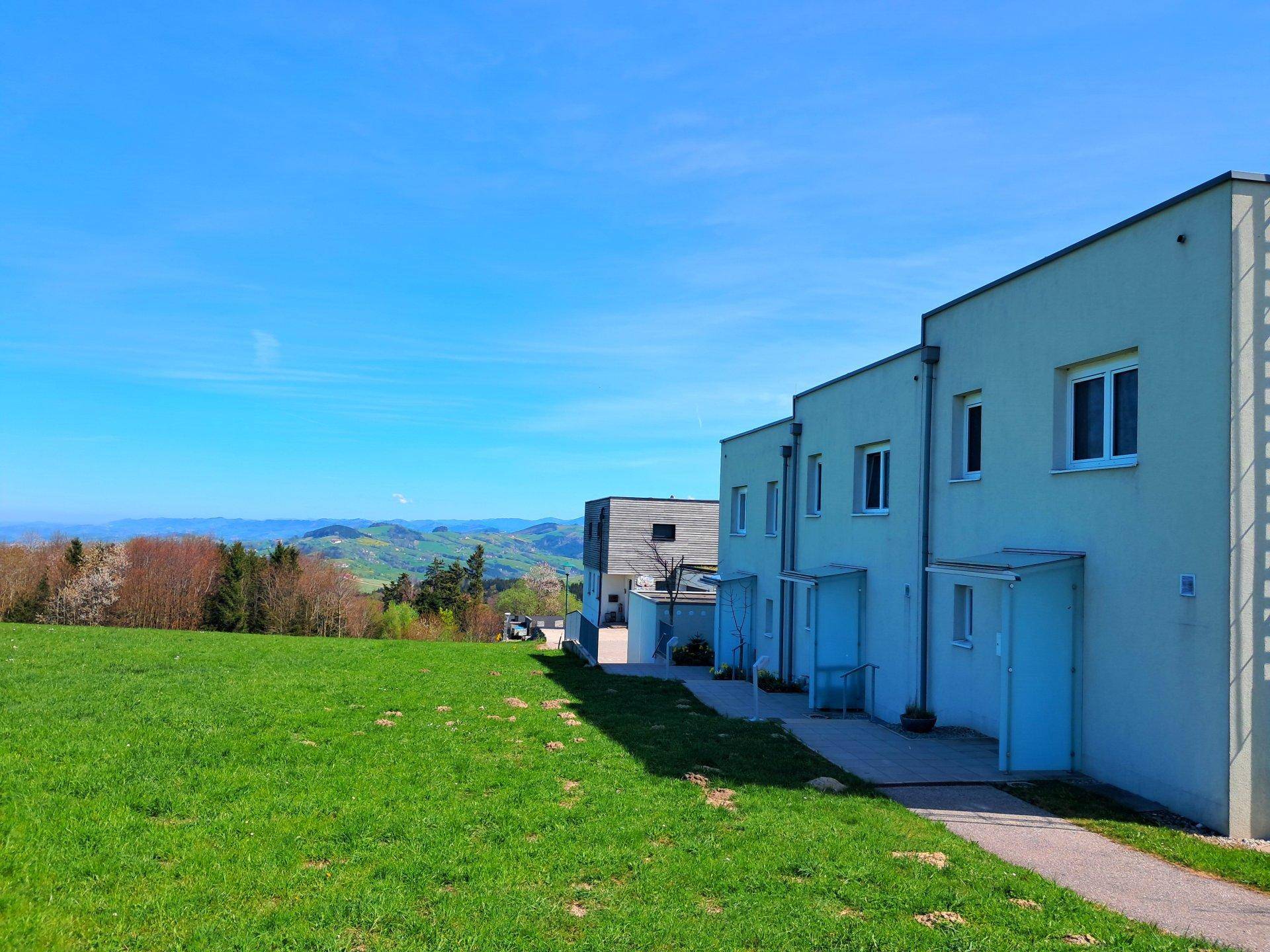 Moderne Wohngebäude mit weitem Blick über grüne Felder und hügelige Landschaft unter blauem Himmel.