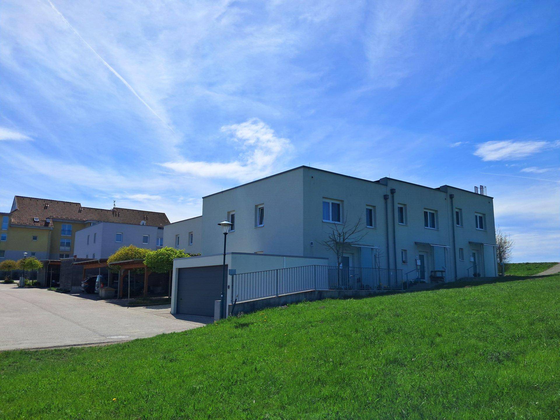 Wohngebäude mit Garage und Carports, angrenzend an eine grüne Wiese unter blauem Himmel.