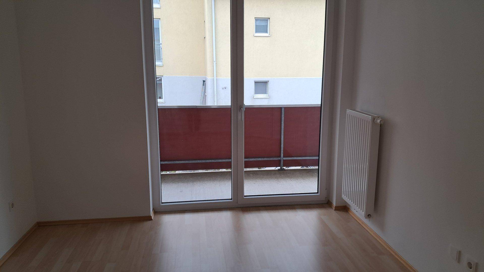 Helles Zimmer mit Laminatboden und großem Balkonfenster, das auf ein gegenüberliegendes Gebäude blickt.