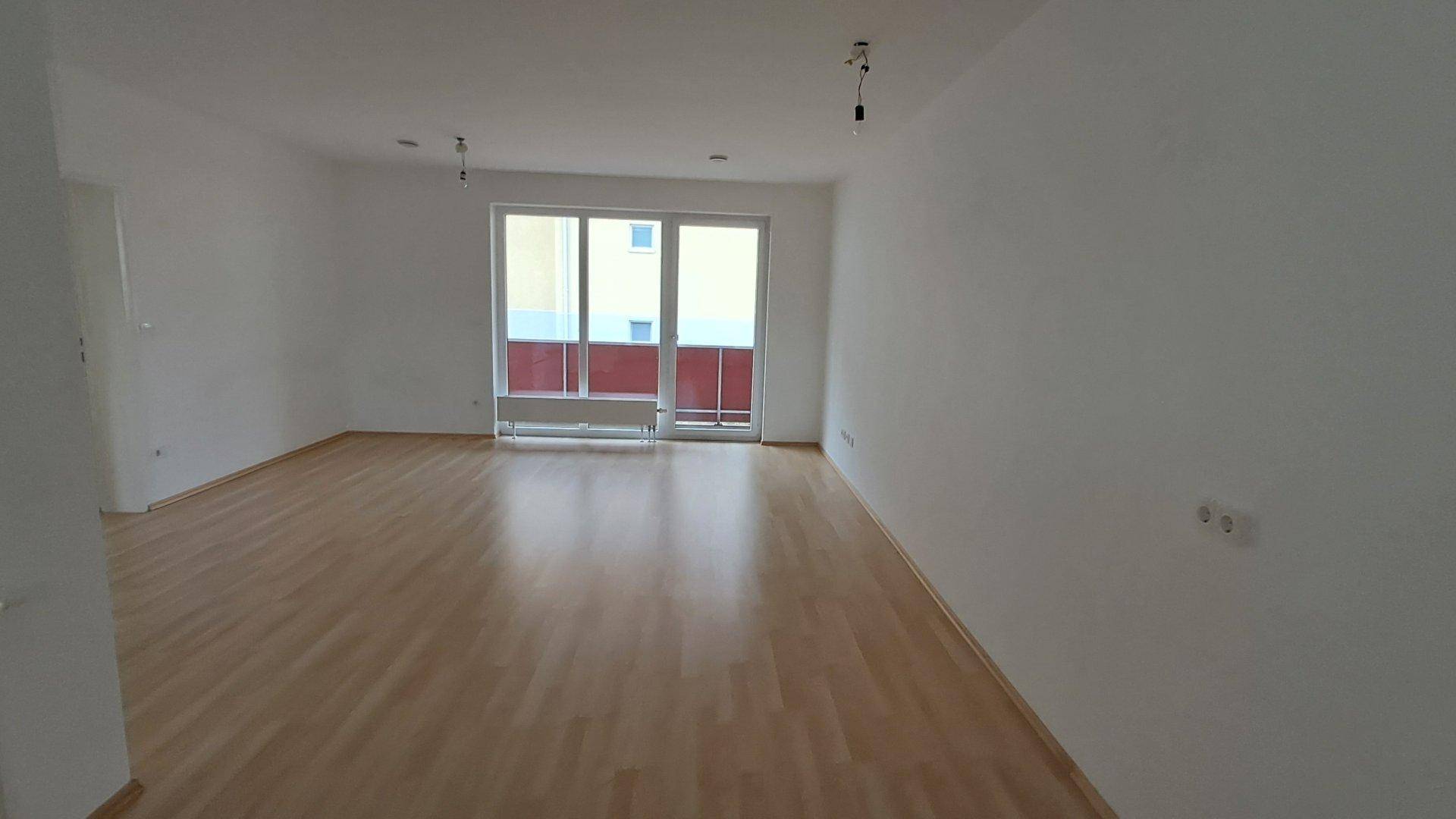 Geräumiges, leeres Wohnzimmer mit hellem Laminatboden und großem Fenster mit Balkonzugang.