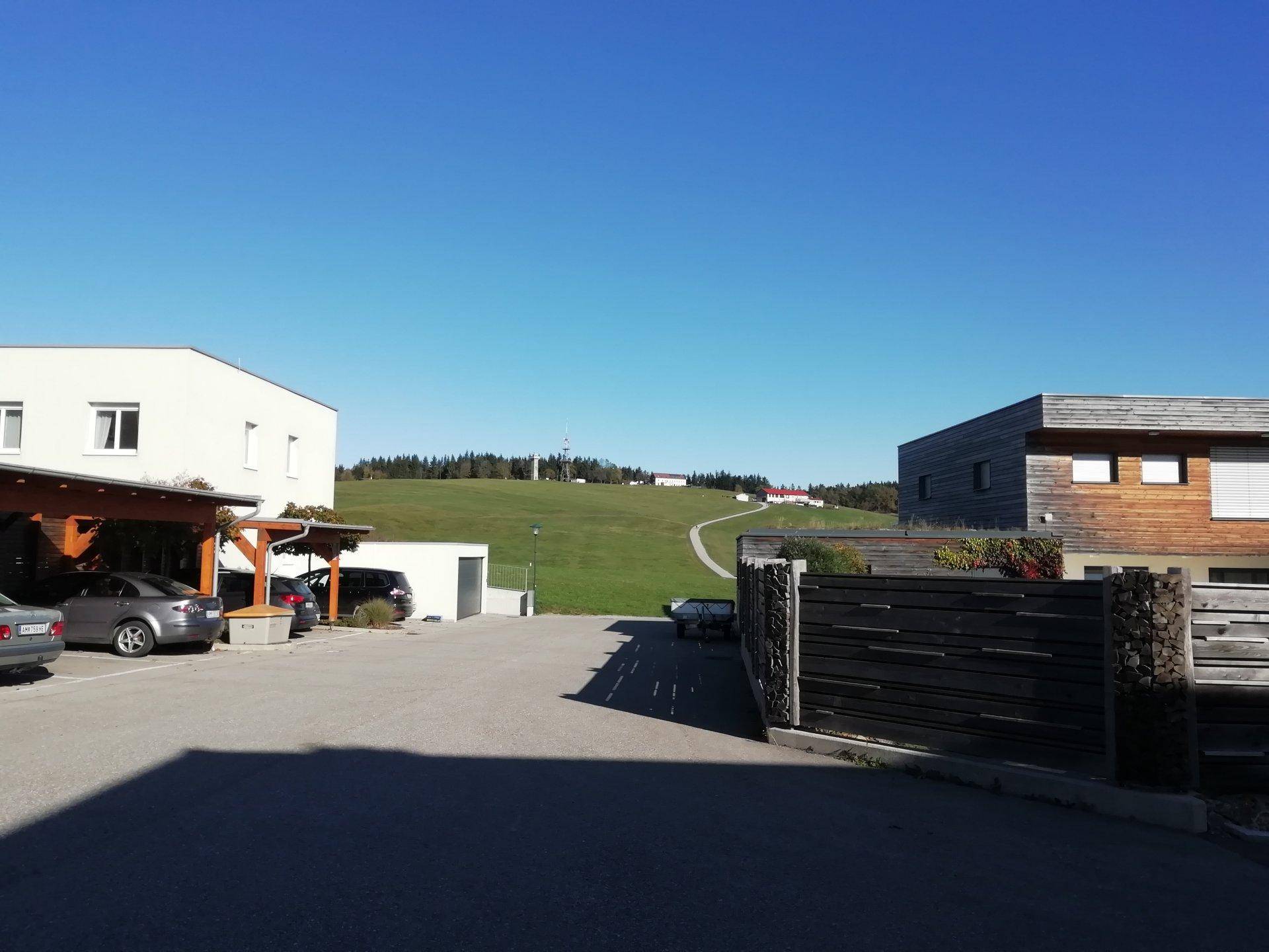 Außenansicht des Anwesens mit Parkplätzen, Carport und weitem Blick auf grüne Hügel unter blauem Himmel.