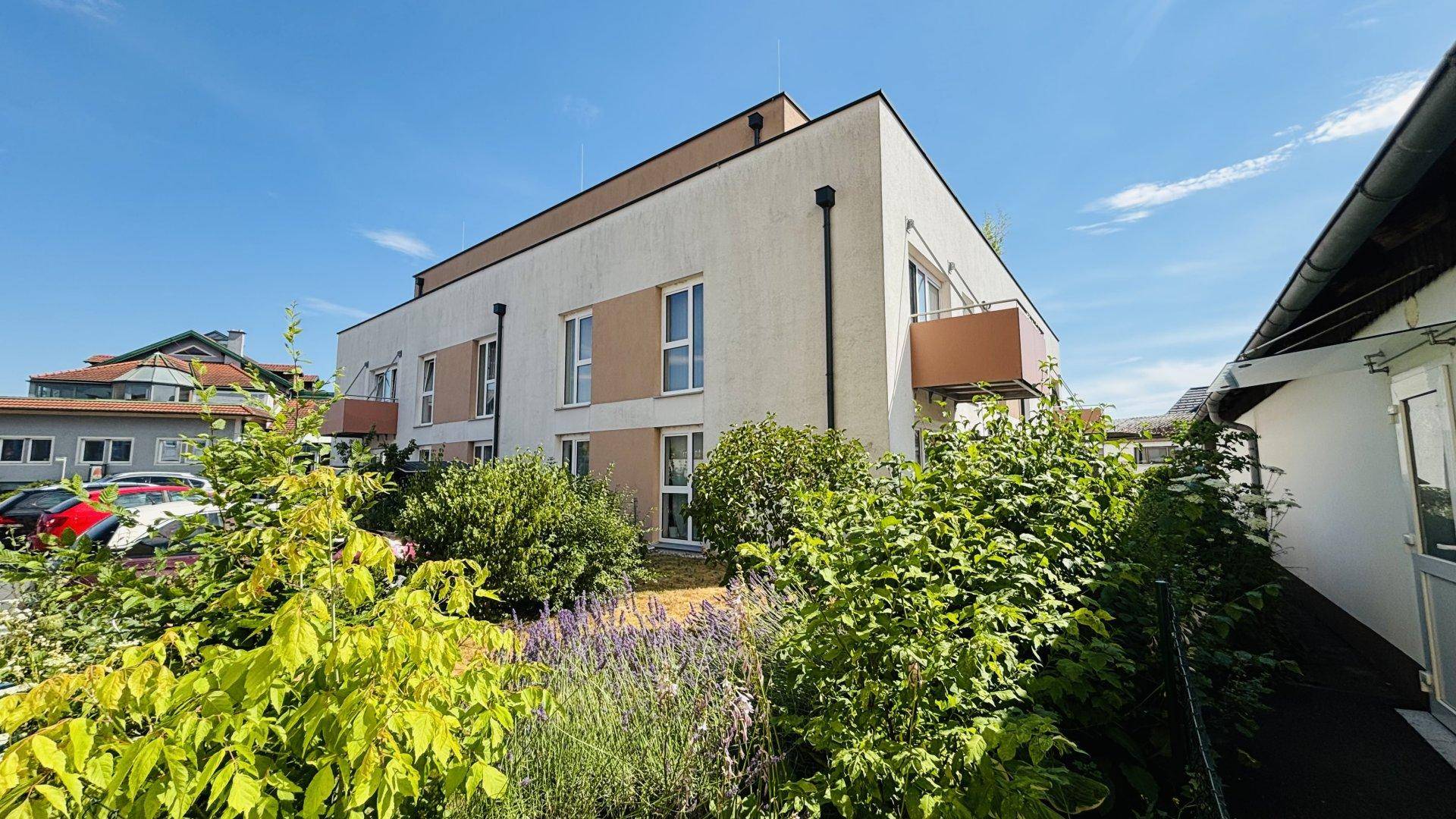 Modernes Mehrfamilienhaus mit heller Fassade, Balkon und gepflegter Bepflanzung im Vordergrund.