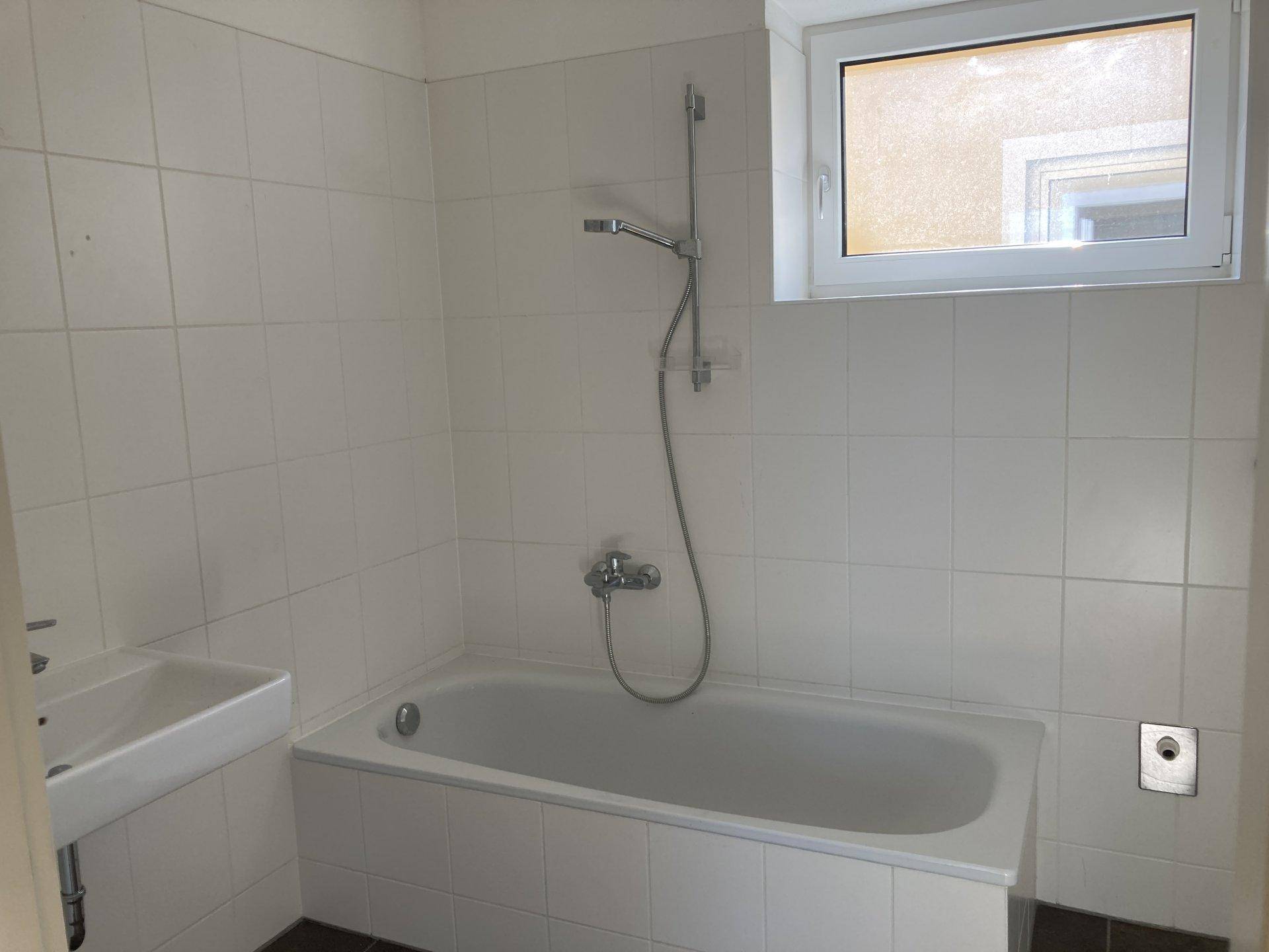 Badezimmer mit weißen Fliesen, Badewanne mit Duschfunktion und einem Fenster für Tageslicht.