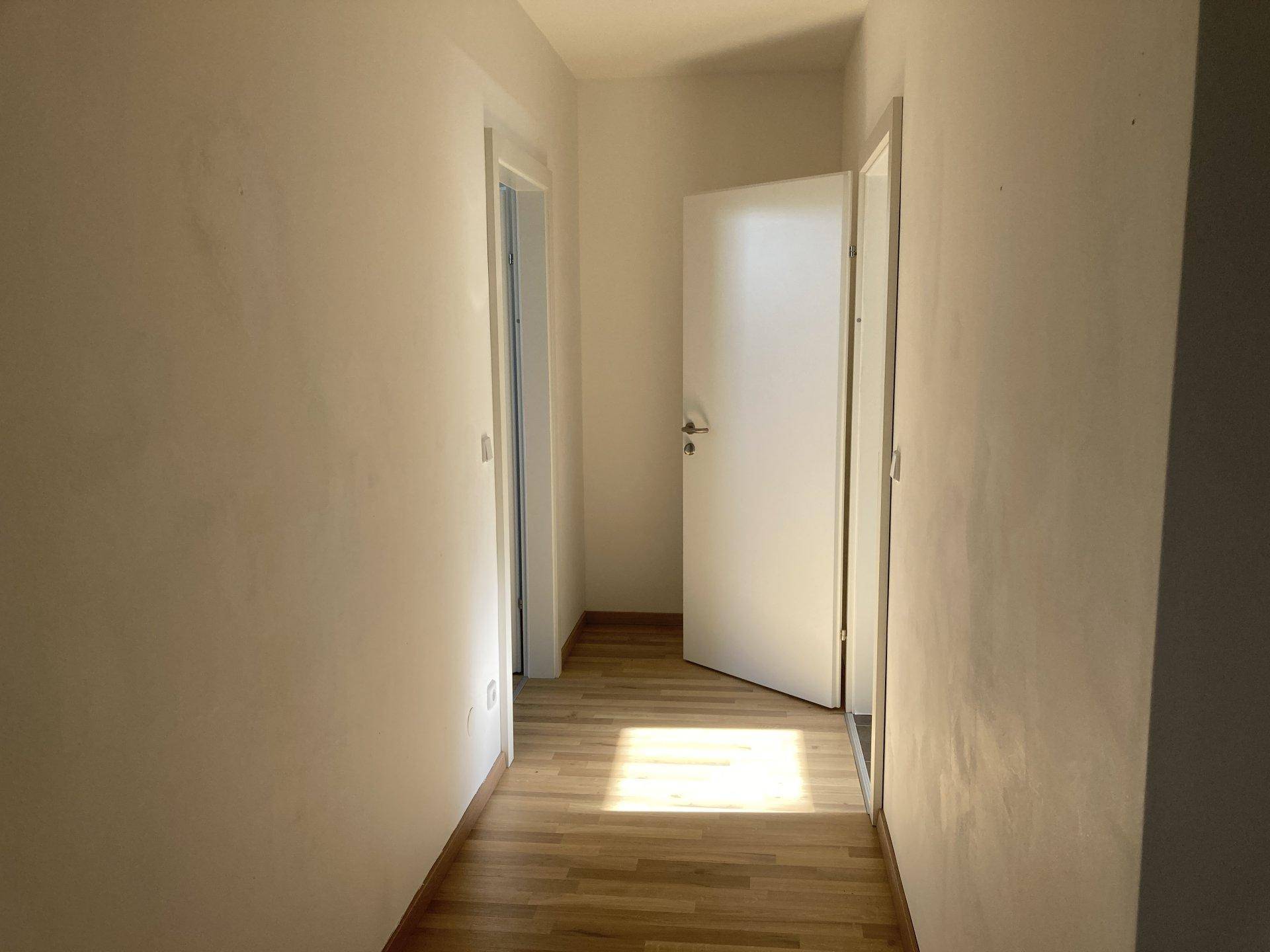 Lichtdurchfluteter Flur mit Holzboden und weiß gestrichenen Wänden, mit Sonneneinfall.