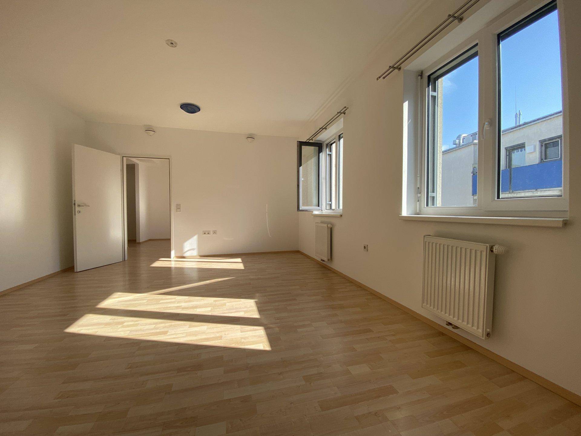 Geräumiges Wohnzimmer mit hellem Laminatboden und großen Fenstern, die viel Sonnenlicht hereinlassen.