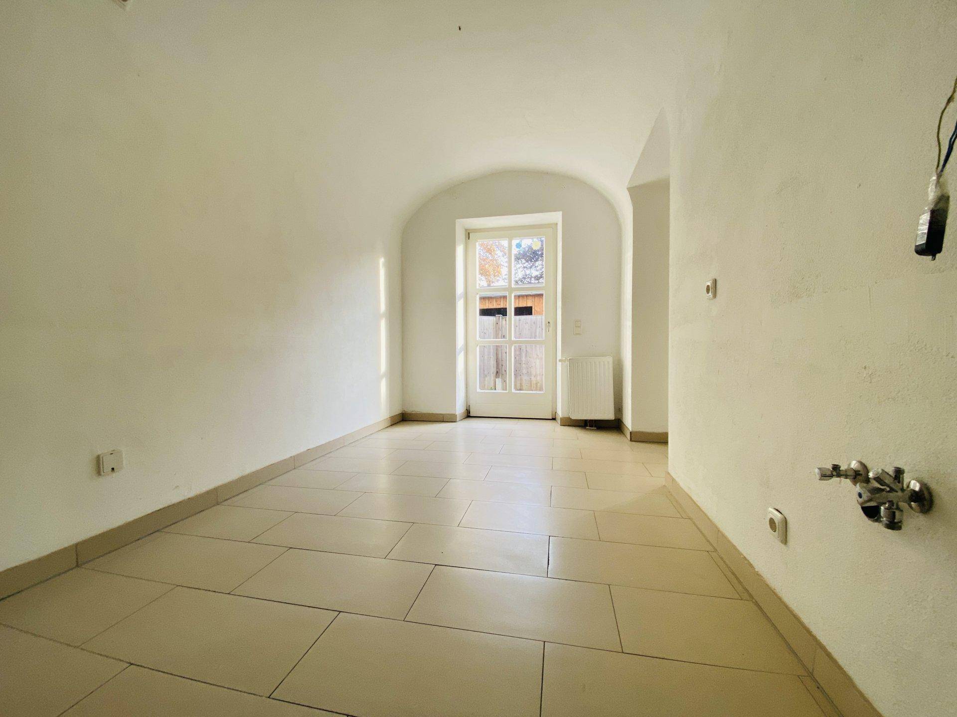 Heller Raum mit Fliesenboden, gewölbter Decke und einer Tür, die ins Freie führt.