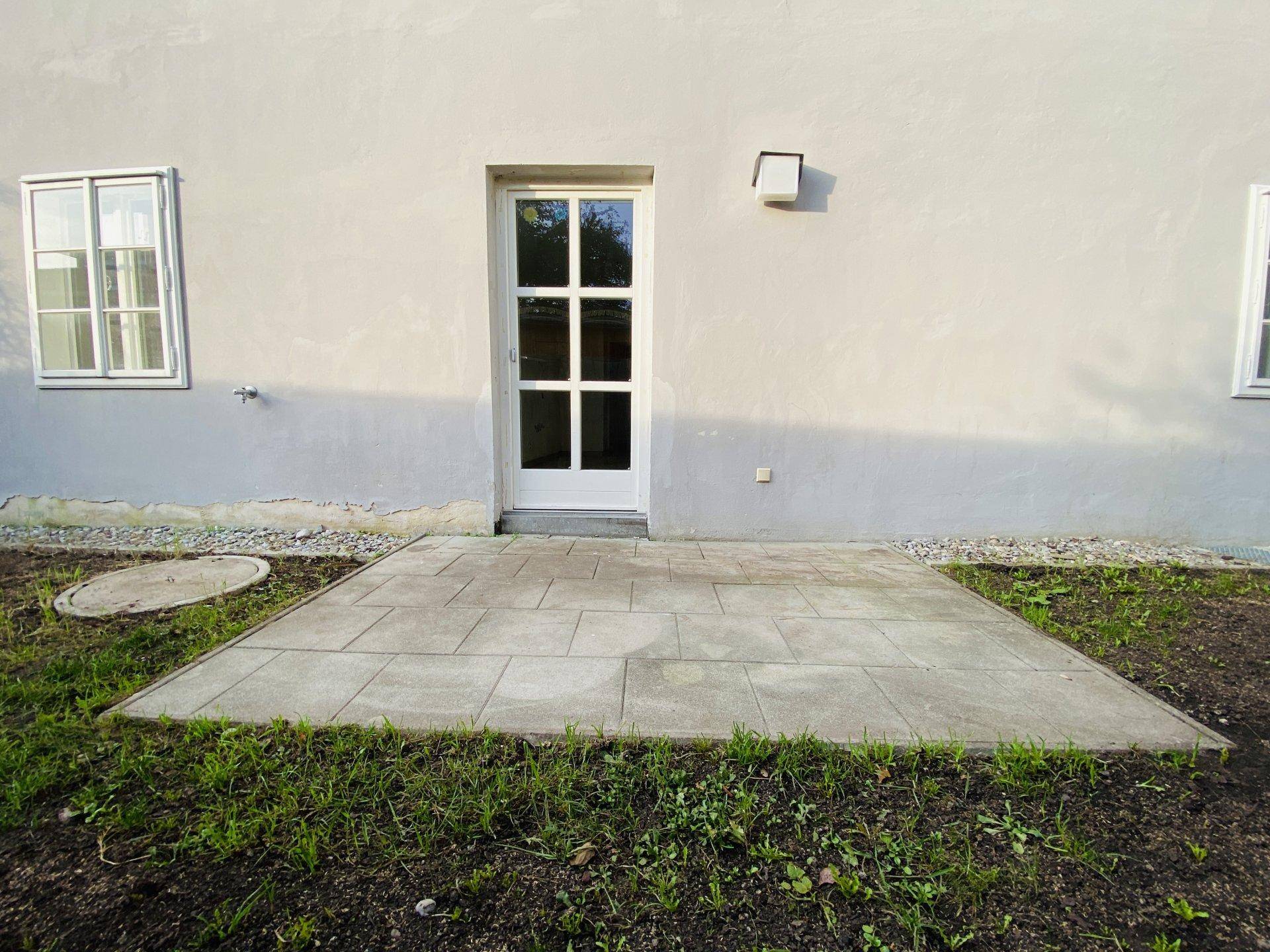 Kleine gepflasterte Terrasse direkt am Gebäude mit Zugangstür und angrenzender Grünfläche.