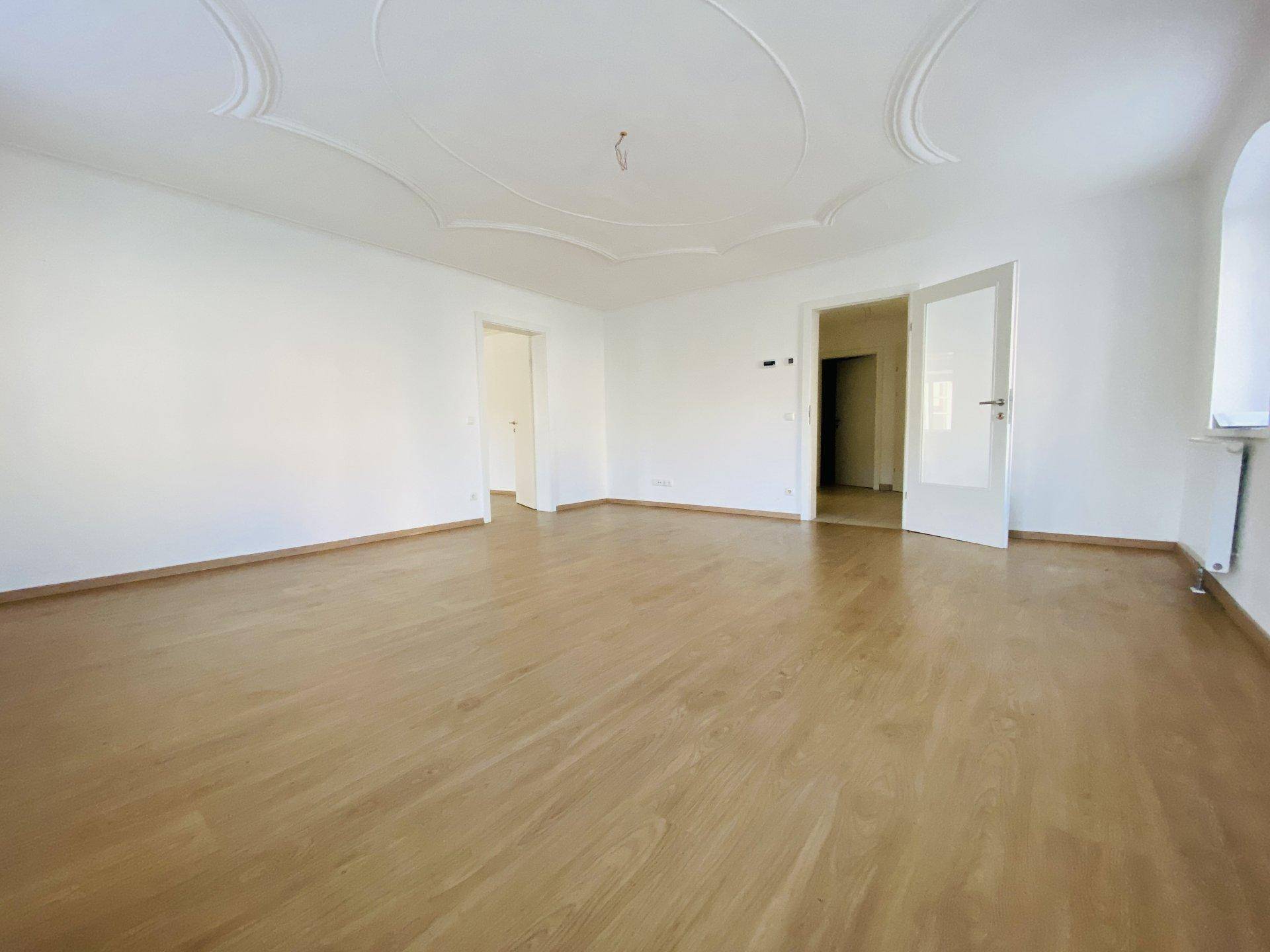 Geräumiges Wohnzimmer mit hellem Holzboden, hohen Decken und dekorativer Stuckverzierung.