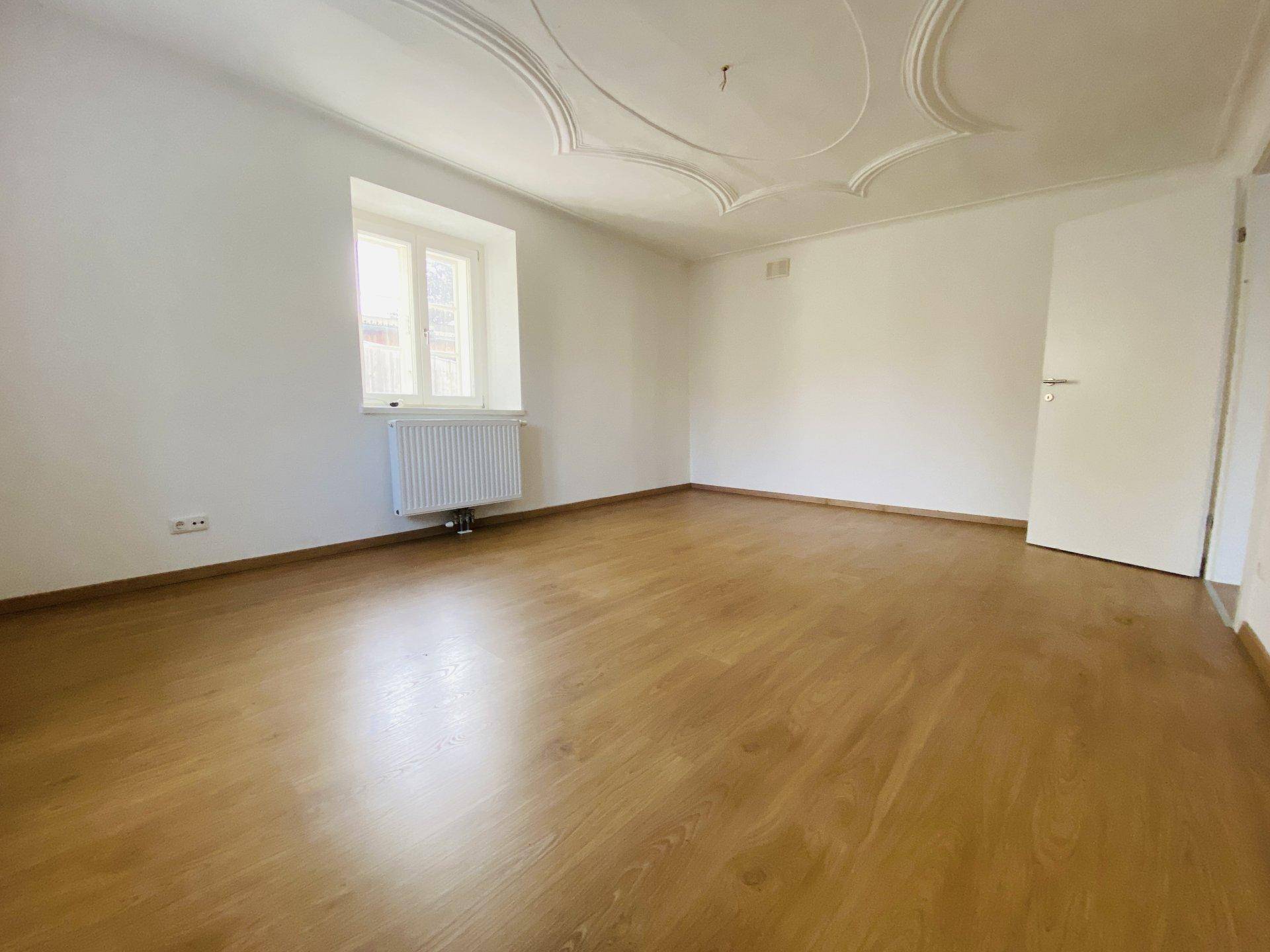 Helles Zimmer mit Holzboden, großem Fenster und eleganter Stuckdecke, bereit zur Einrichtung.