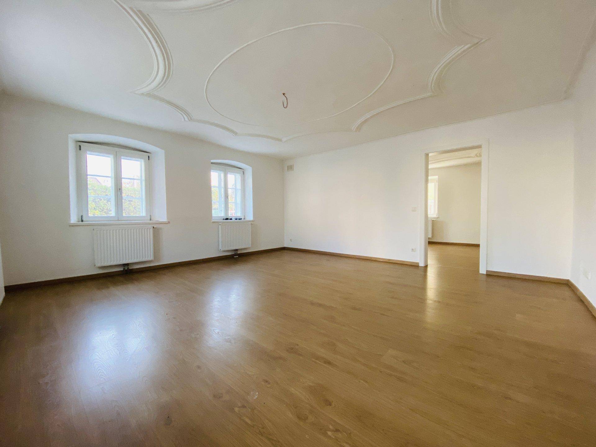 Großes, lichtdurchflutetes Zimmer mit Holzboden, zwei Fenstern und kunstvoller Stuckdecke.
