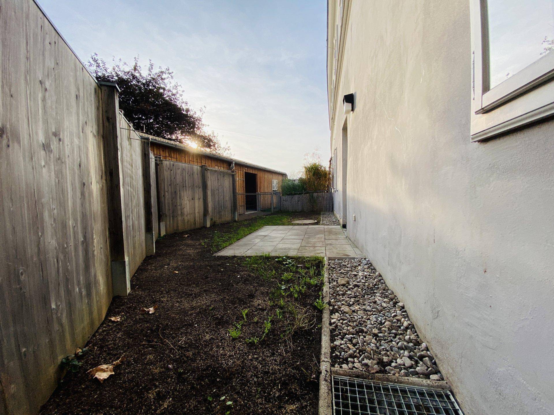 Seitlicher Gartenweg mit Kiesstreifen, Holzzaun und einem Schuppen im Hintergrund.