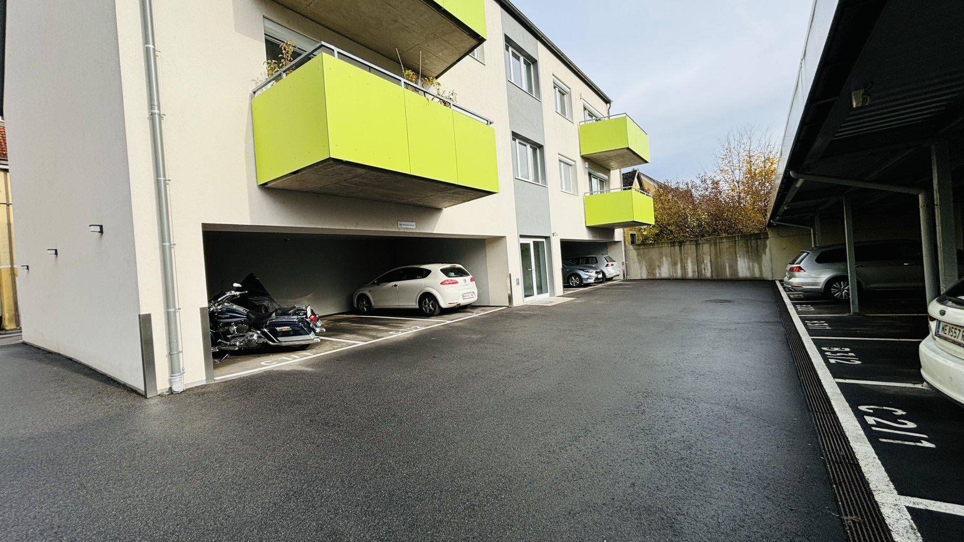 Rückseite des Gebäudes mit überdachten Parkplätzen und Balkonen mit grünen Brüstungen.