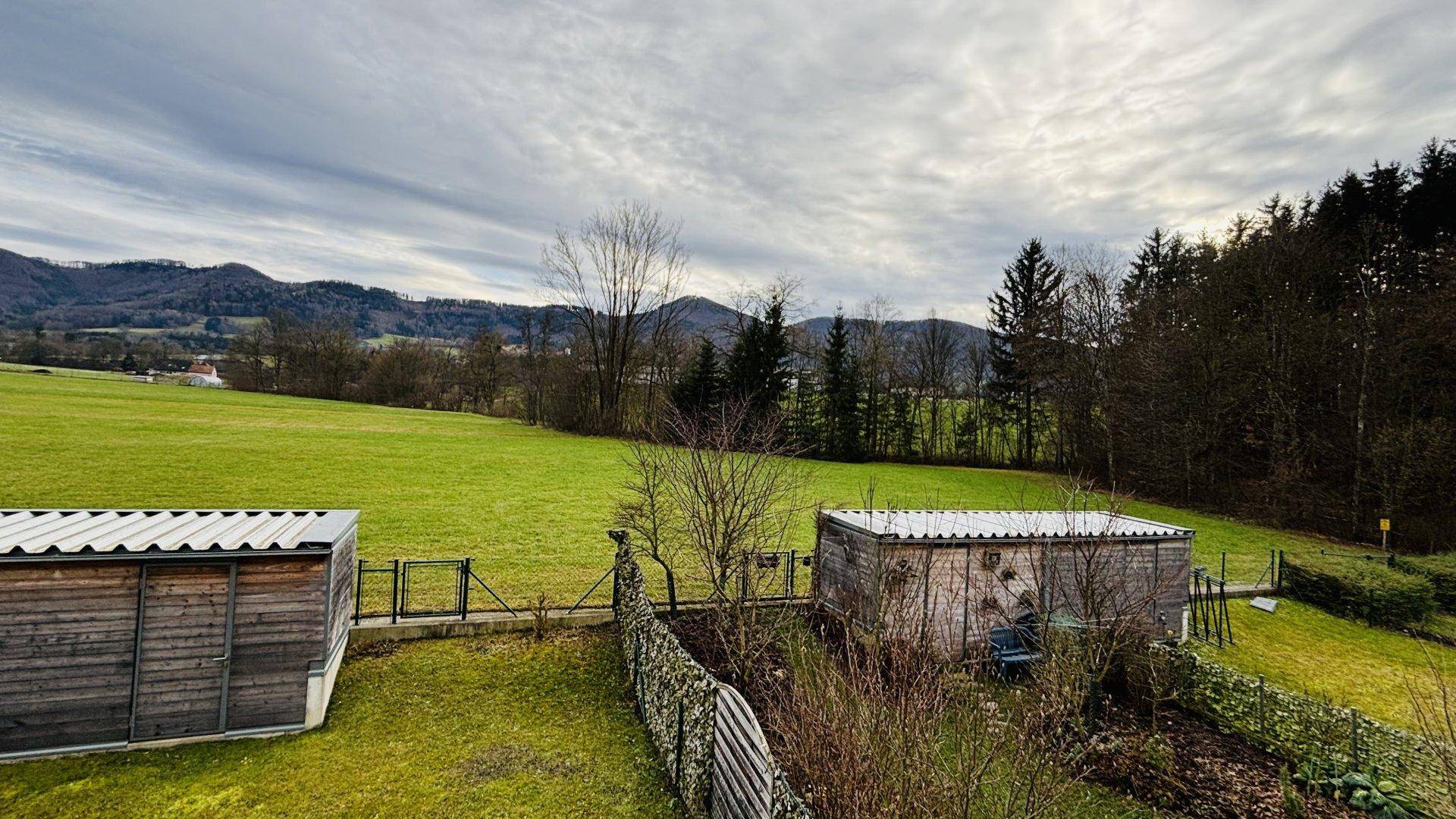 Weitläufiger Blick über grüne Wiesen und bewaldete Hügel unter einem bewölkten Himmel.