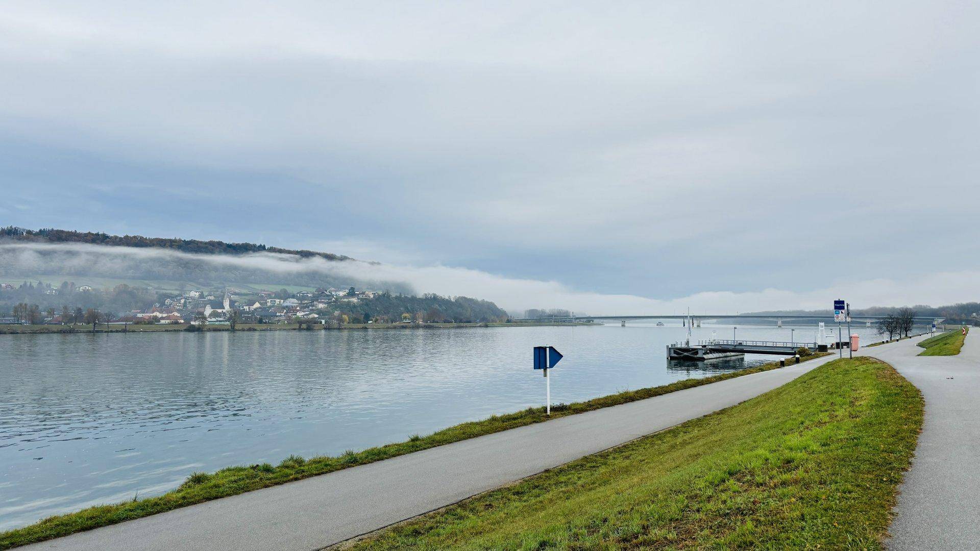 Weitläufiger Blick auf einen Fluss mit Brücke und nebligen Hügeln am Horizont.