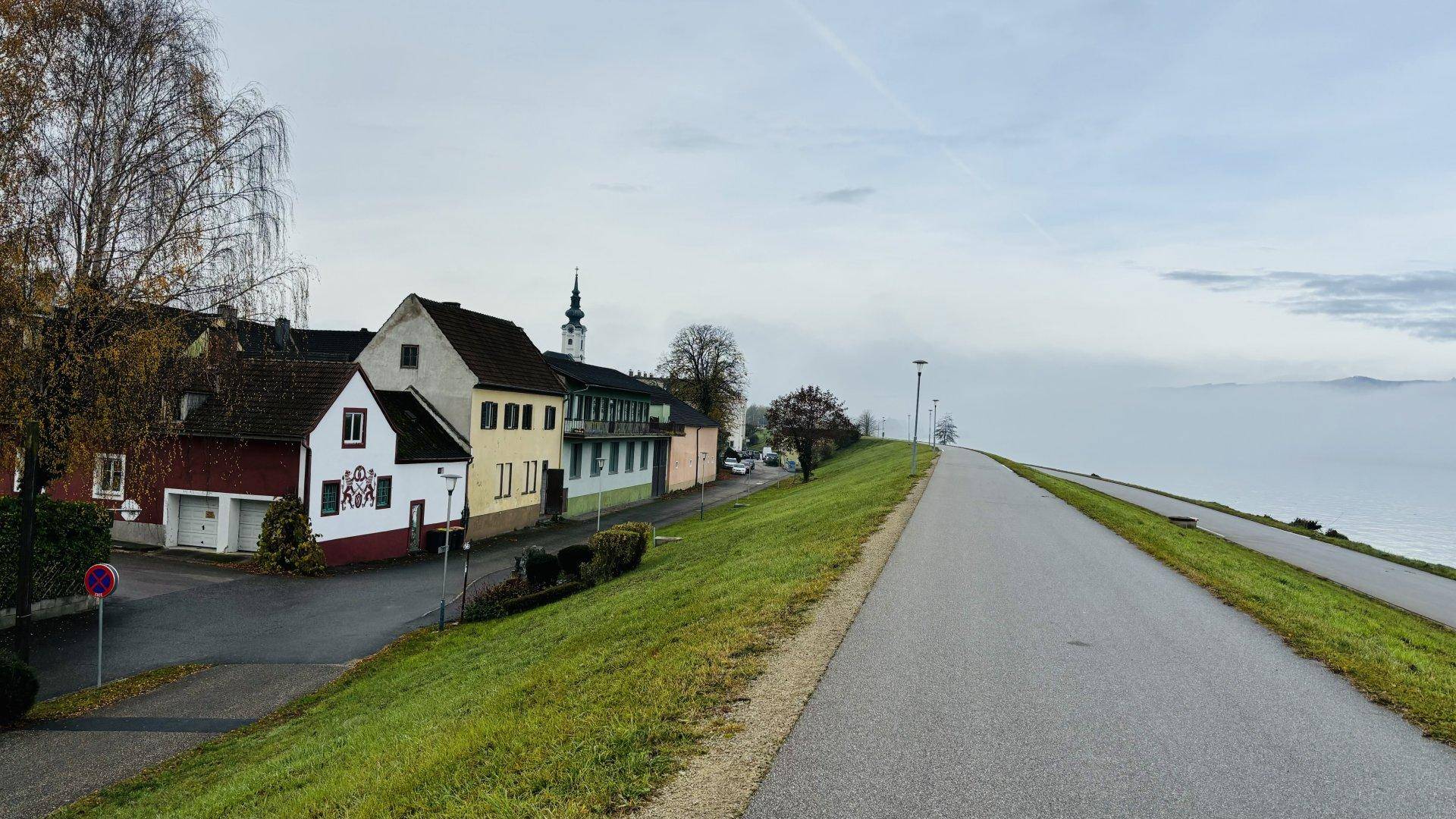 Ländliche Straße entlang eines Gewässers mit Häusern und bewölktem Himmel.