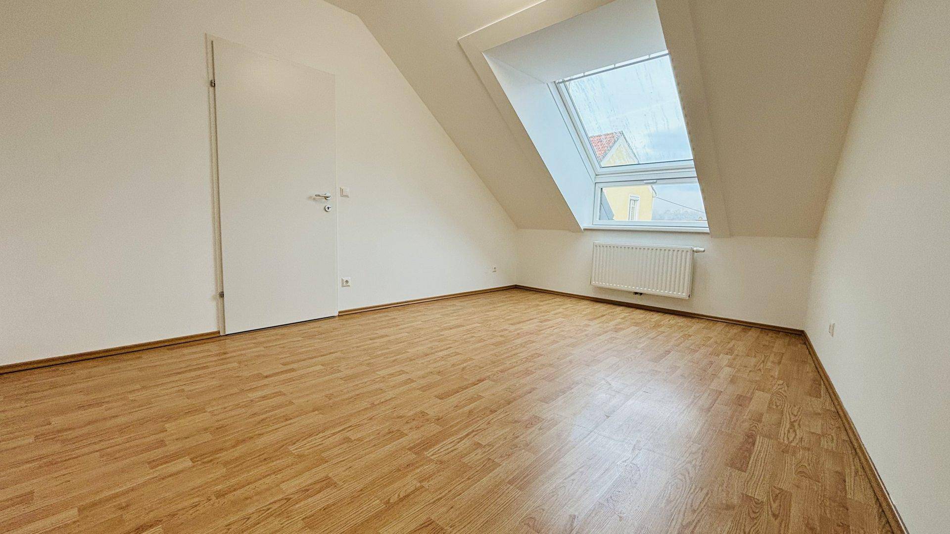 Helles Zimmer im Dachgeschoss mit Laminatboden, Dachfenster und schrägen Wänden.