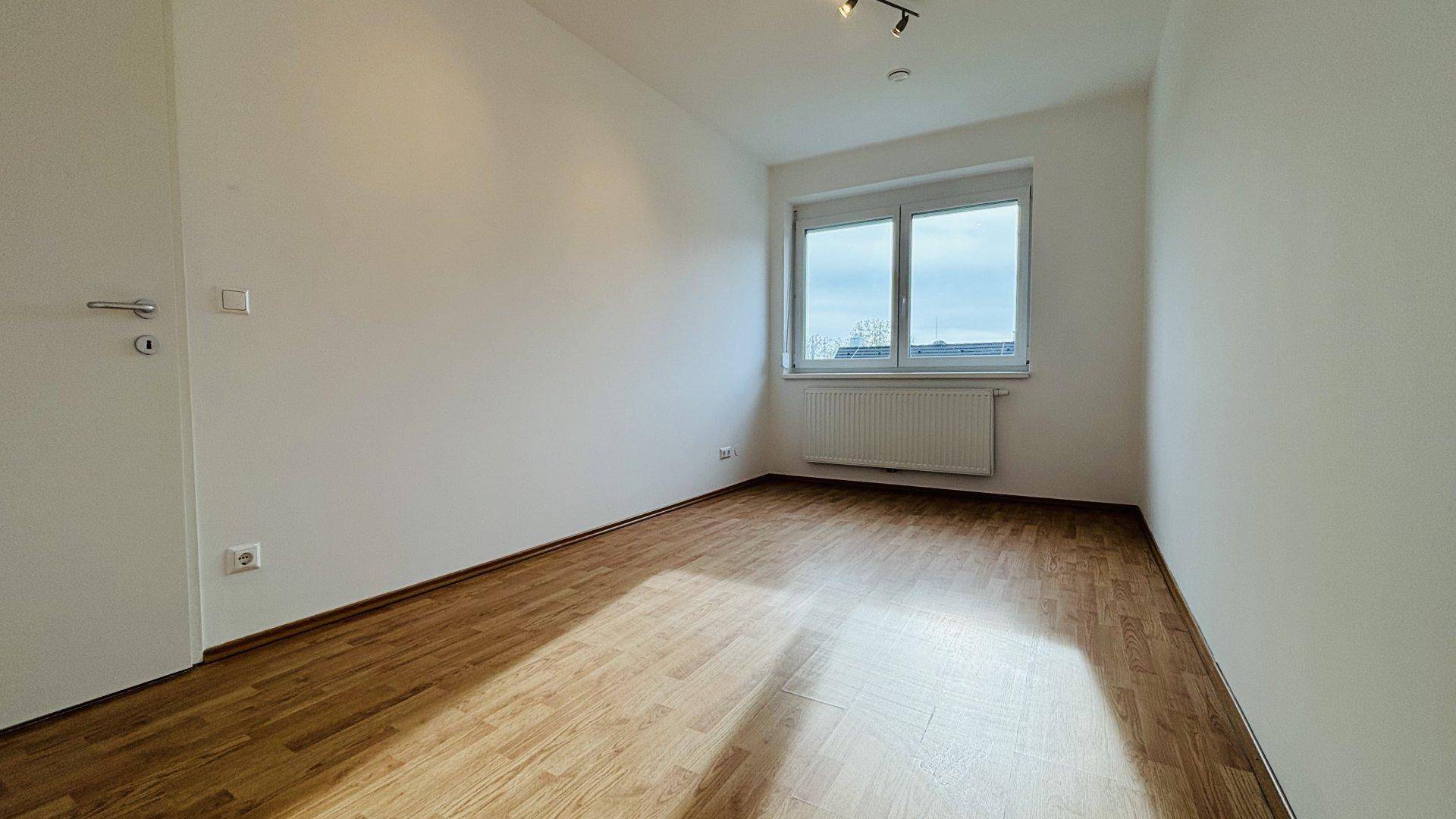 Leeres Zimmer mit Laminatboden, großem Fenster und Heizkörper unter dem Fenster.
