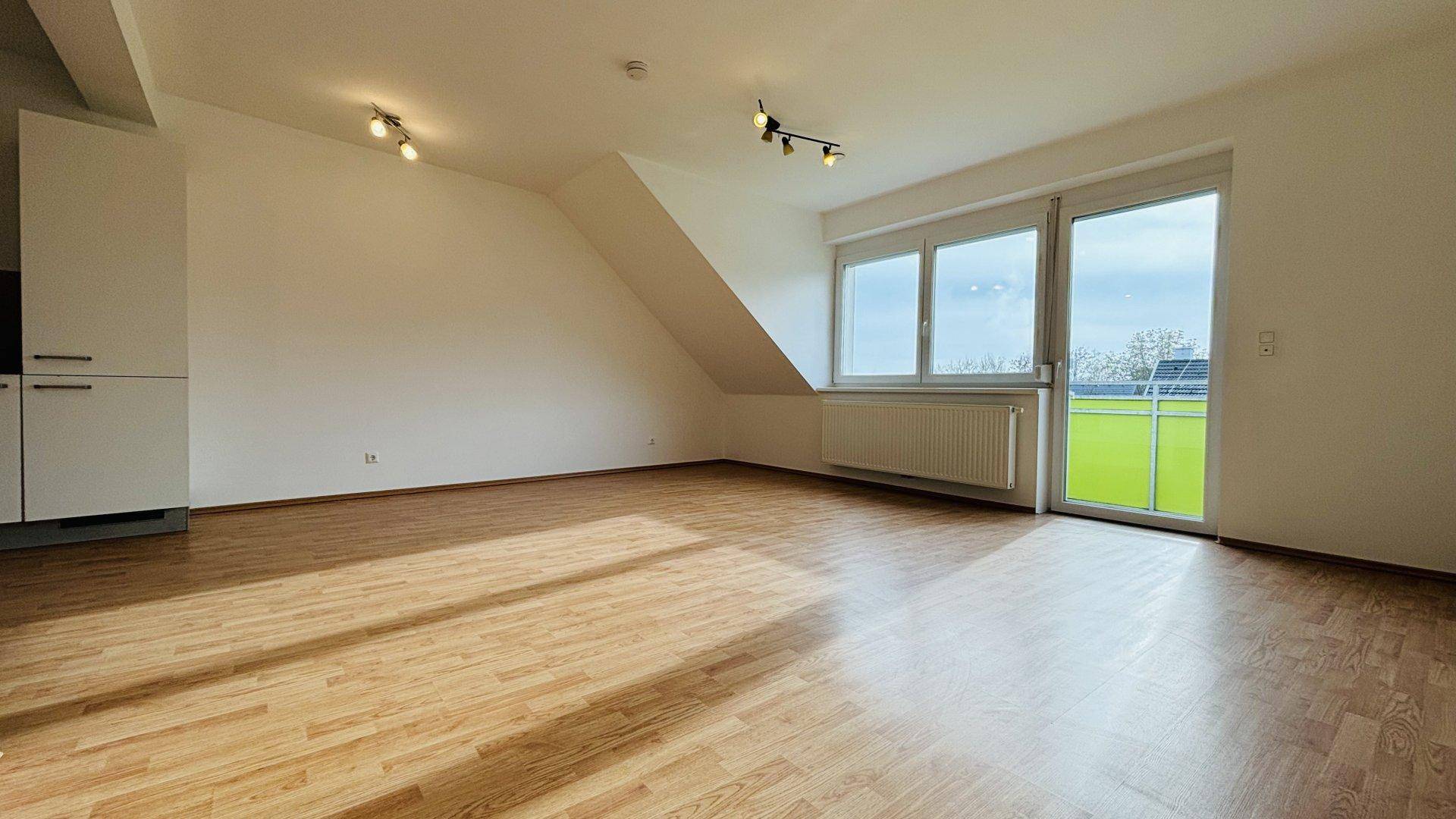 Offener Wohnbereich mit Laminatboden, Zugang zum Balkon und schräger Decke.